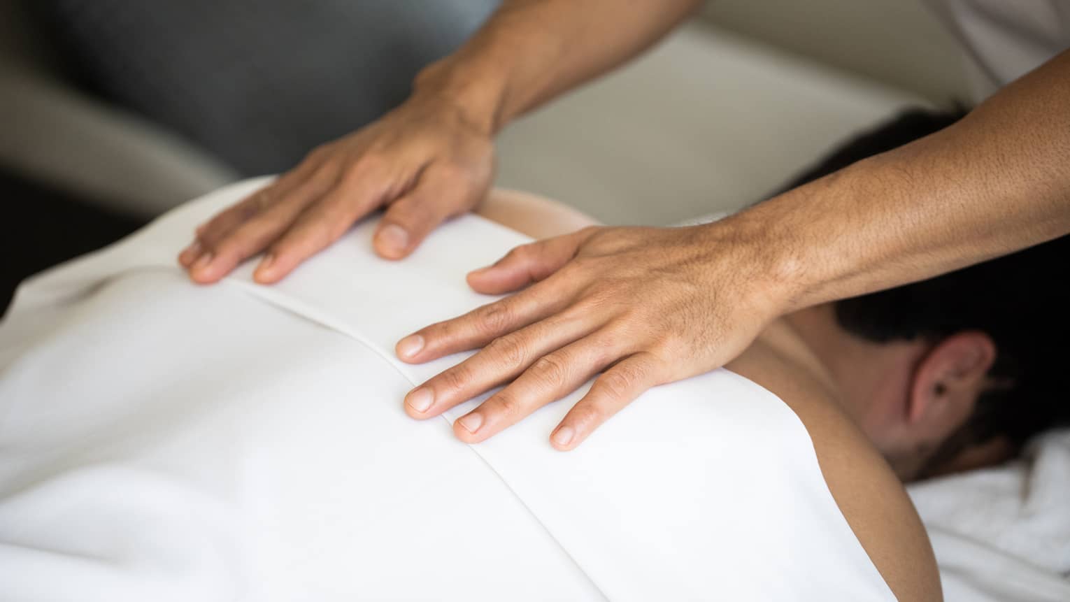 Two hands lay flat on the back of a man laying face-down on a massage table covered with a white sheet