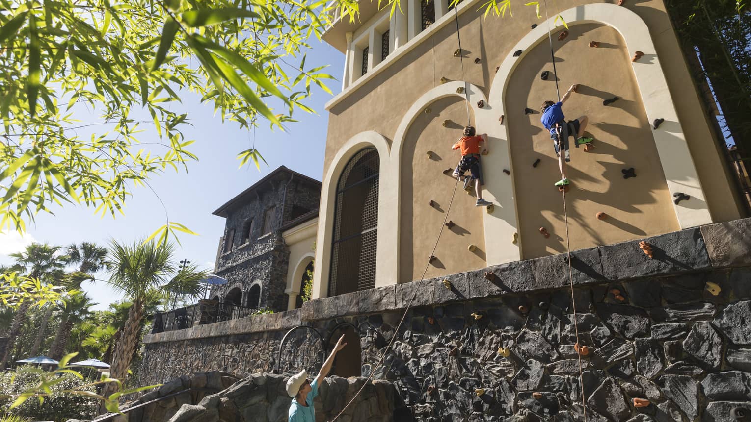 Two people climbing an outdoor rock climbing wall set along the side of a building with an instructor standing on the ground