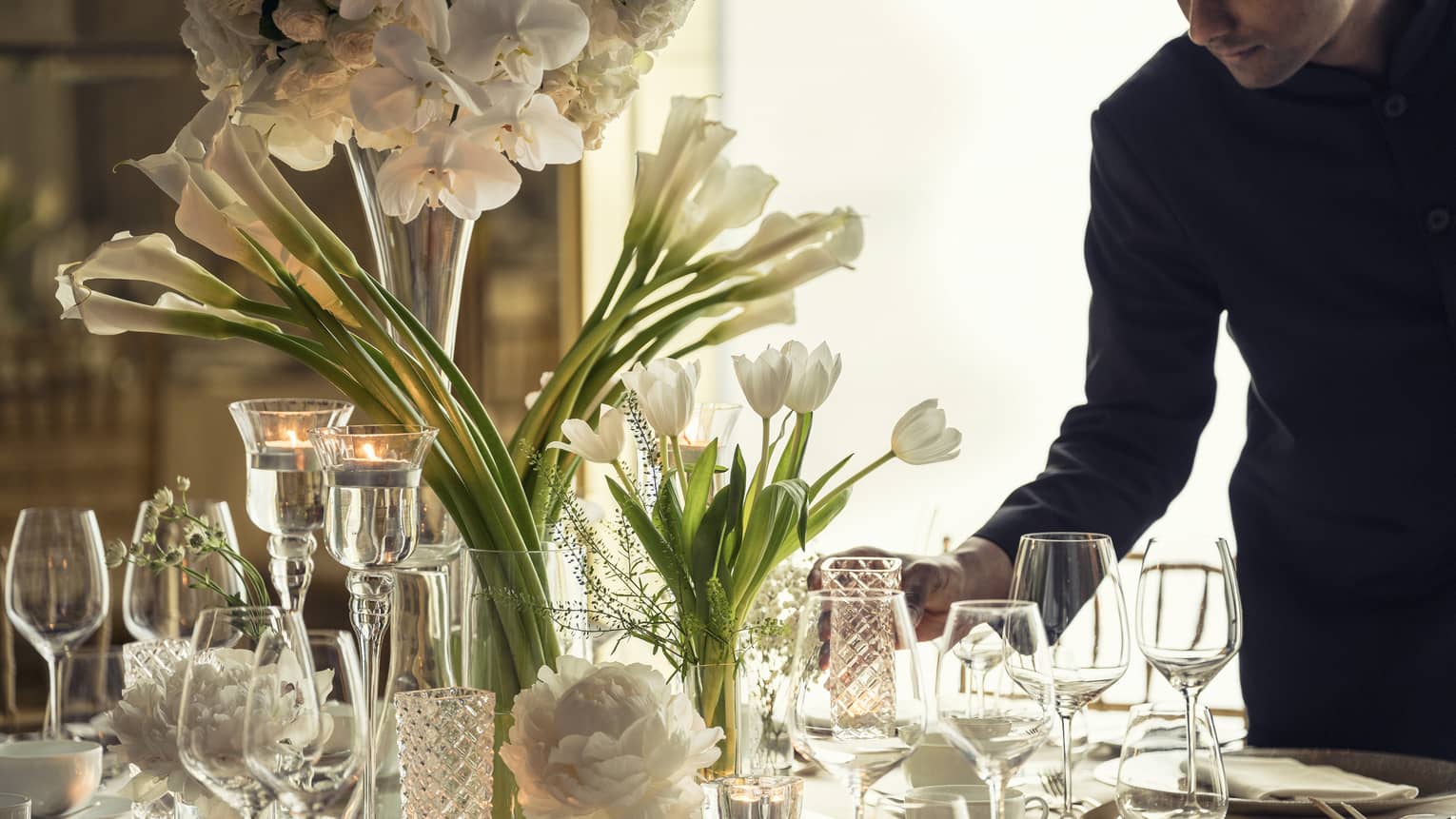 Hotel staff sets elegant banquet dining table with crystal glasses, vases with white flowers