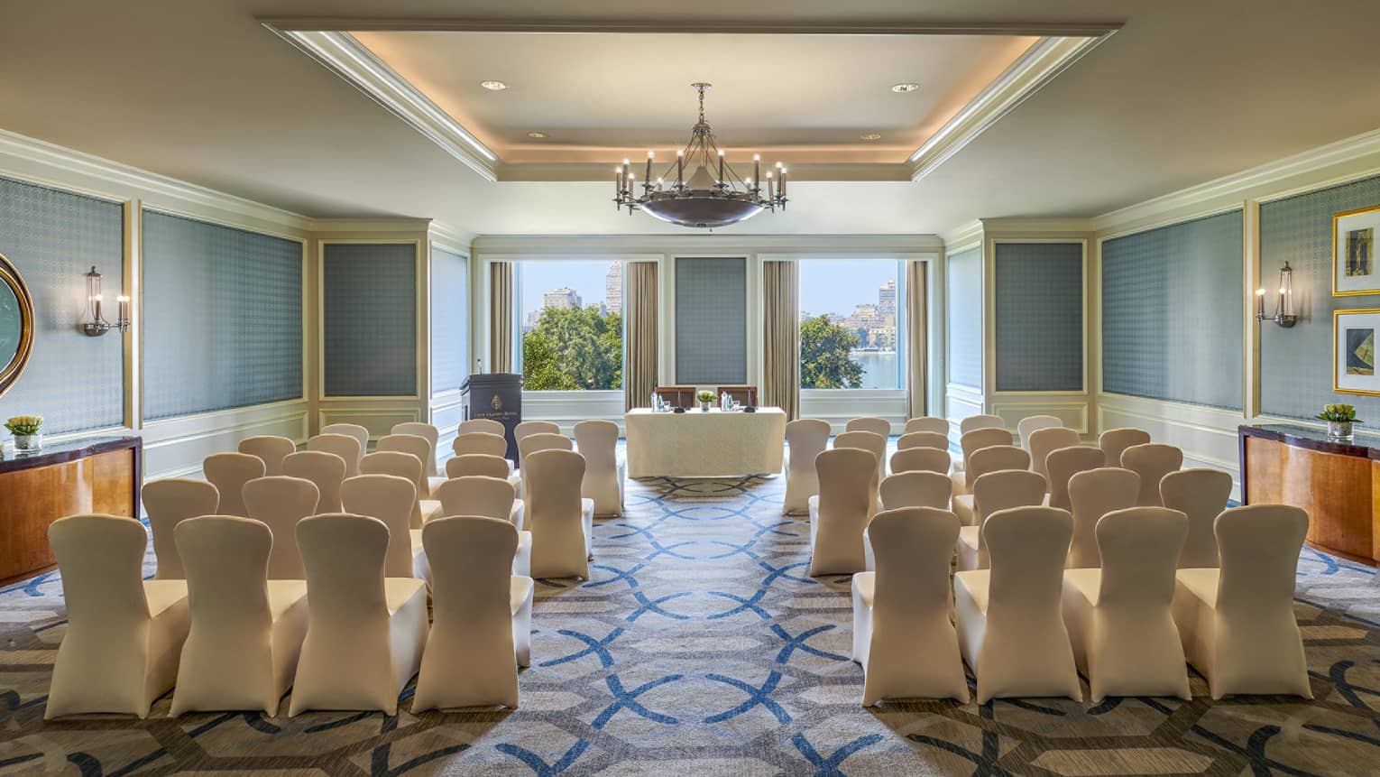 Rows of fabric covered chairs face conference table, podium and sunny windows 