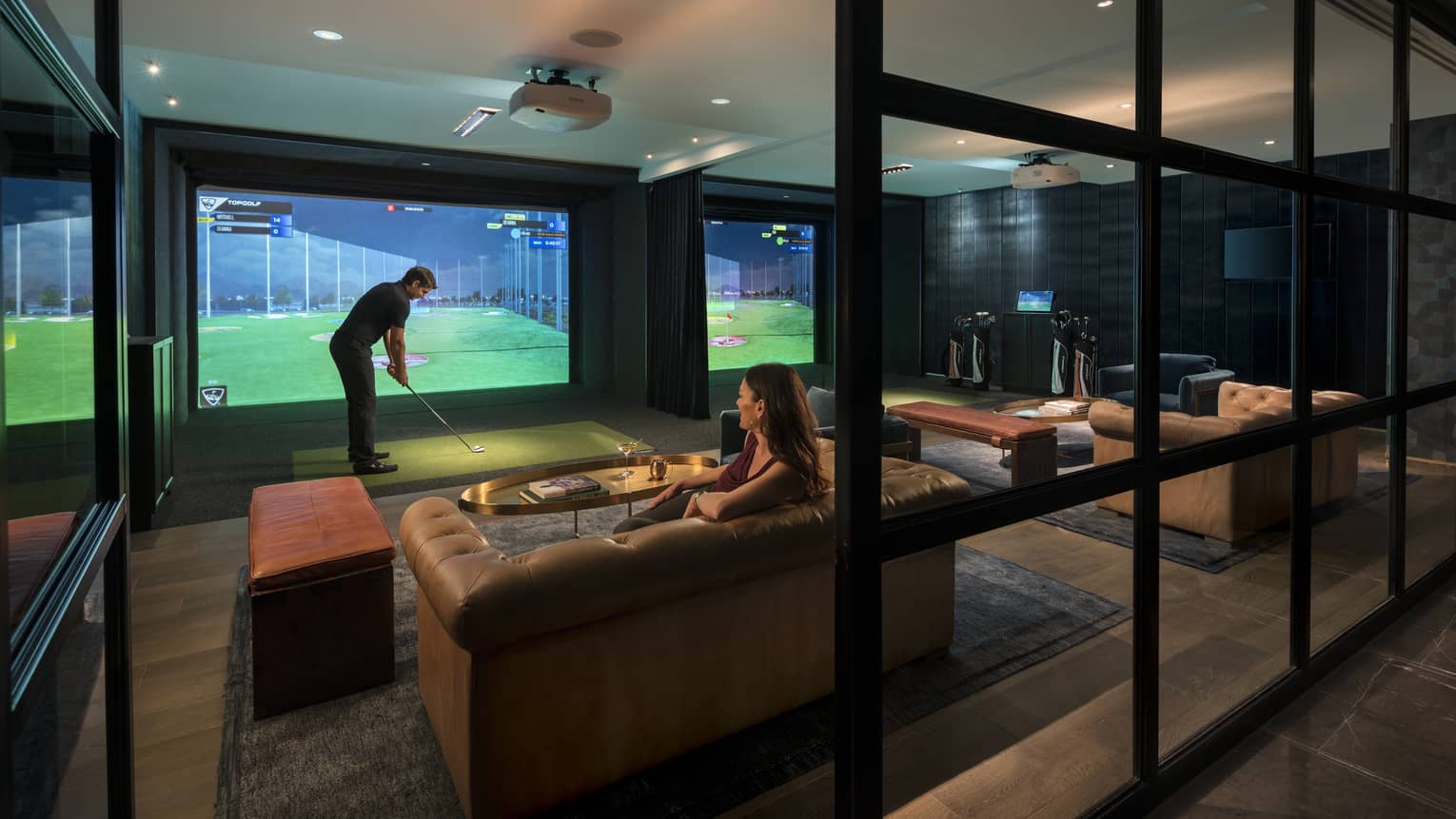 Woman lounges on sofa as man swings golf club in front of screen at Topgolf Swing Suite inside Bayou & Bottle.