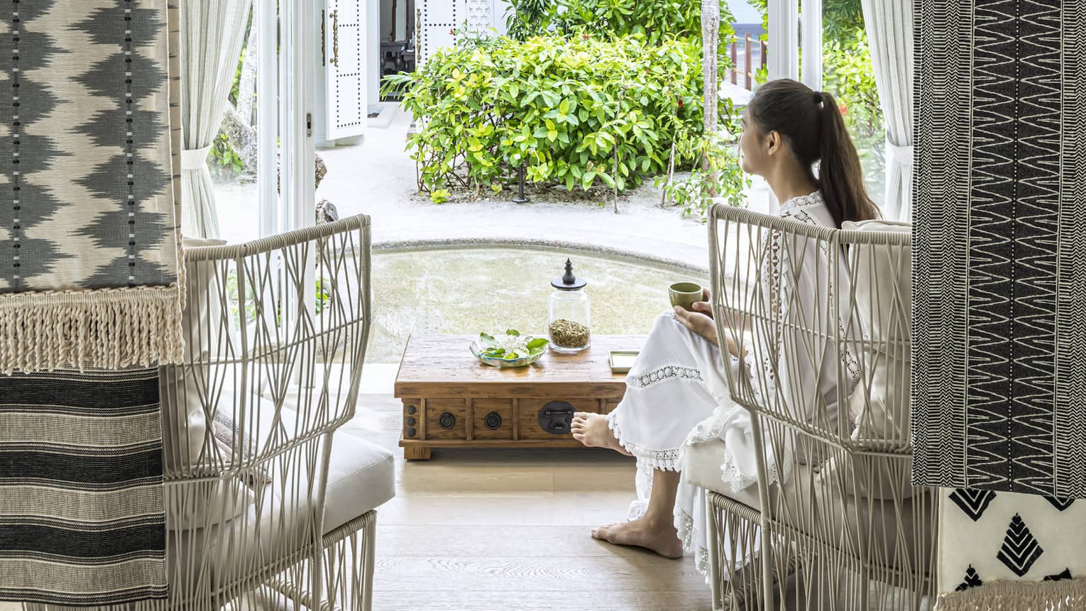 A woman sits in white wicker chair in the four seasons maldives spa waiting area and overlooks the garden outside an opened window