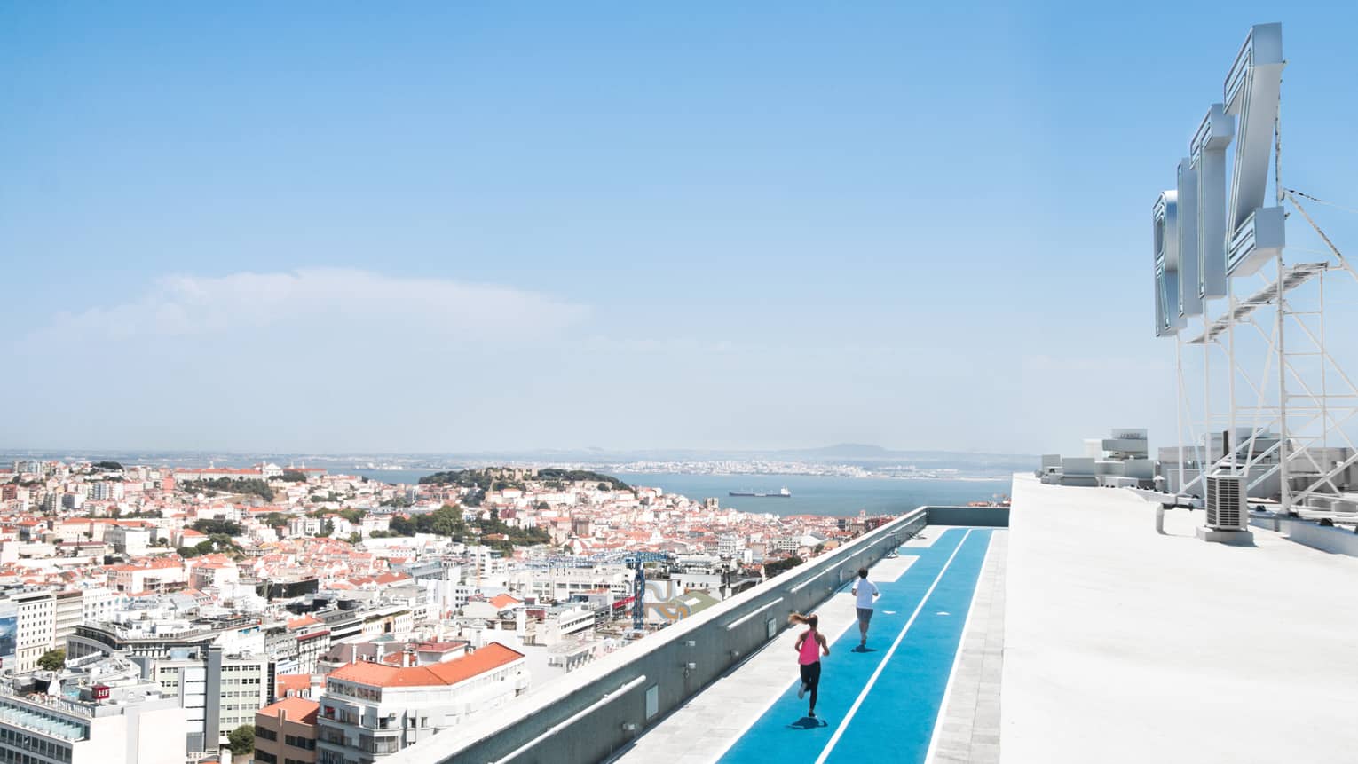 Aerial view of backs of man and woman running on blue rooftop outdoor track, Lisbon city roofs below