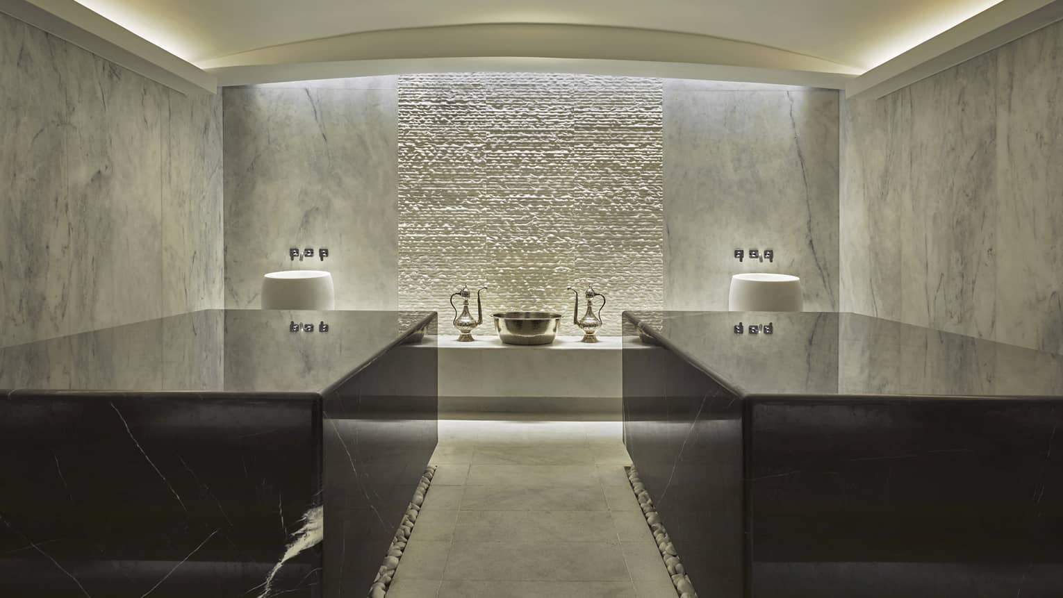 The interior of a hammam with grey marble walls, two black marble tables and a gold sink in the backfround