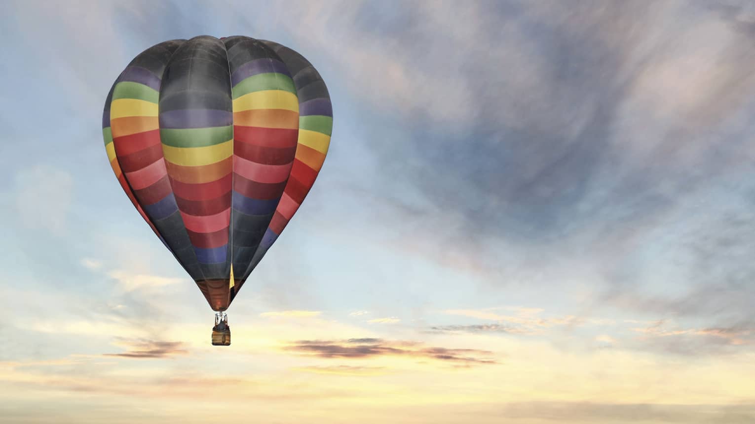 Rainbow-striped hot air balloon floats in the sky at sunset