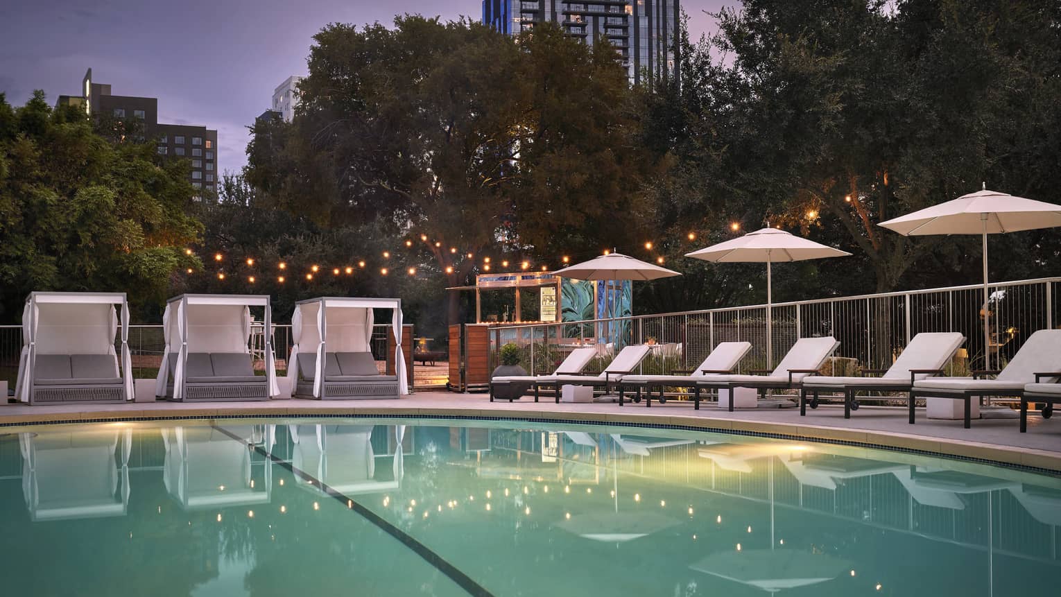An outdoor pool at evening surrounded by lounge chairs, umbrellas and large trees.
