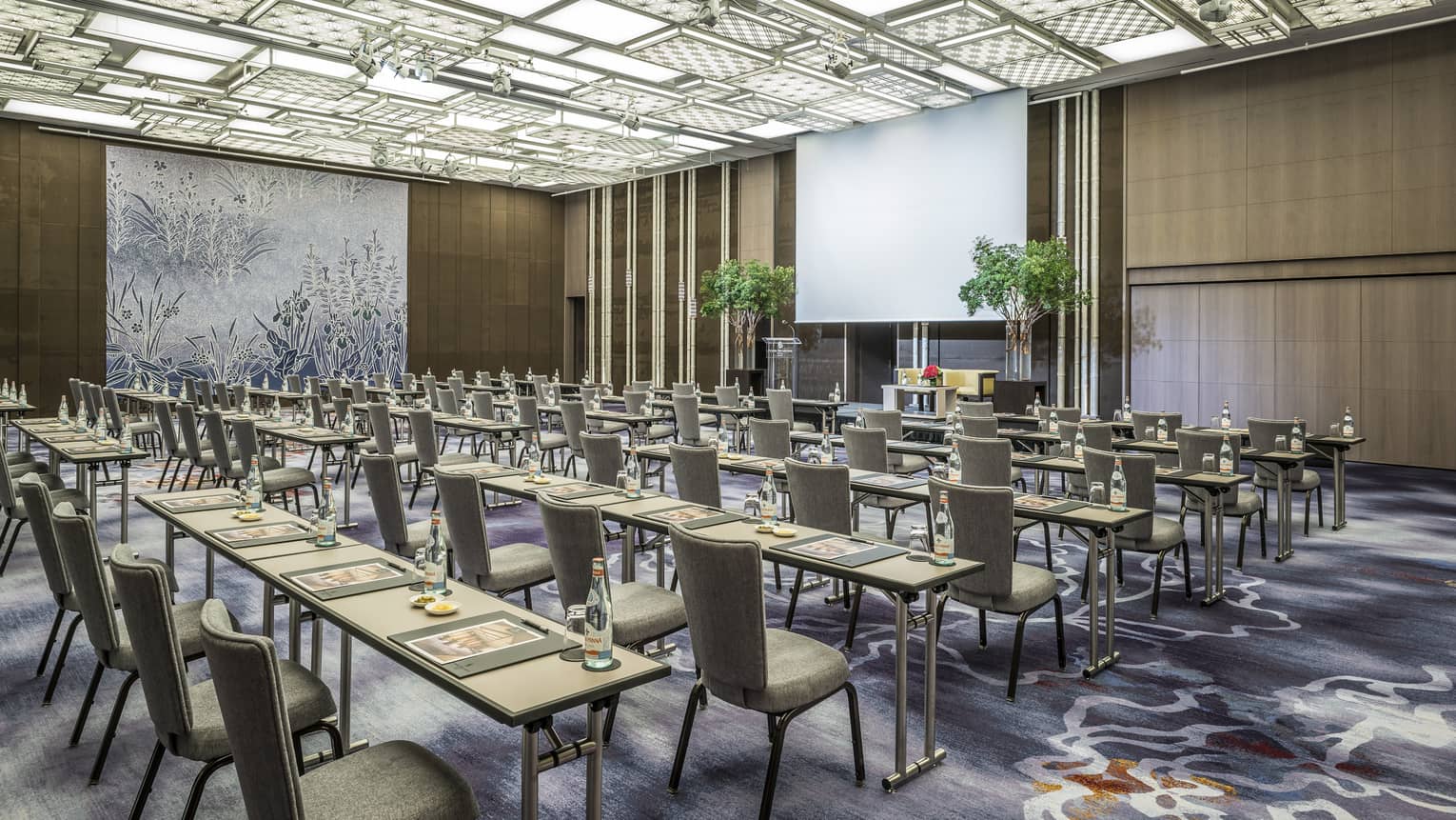 Rows of meeting tables, chairs facing large projector screen in ballroom conference room