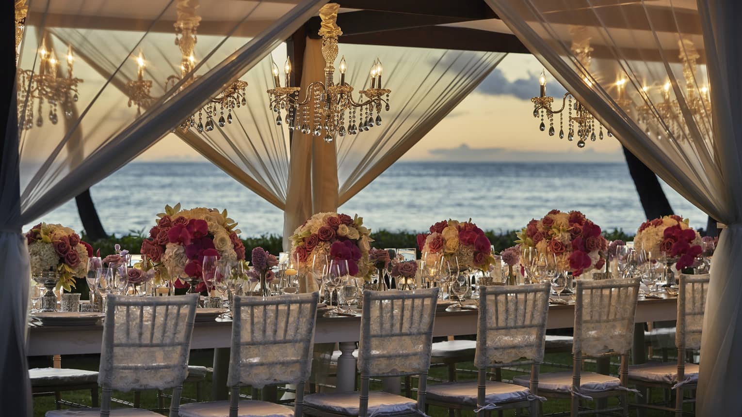 Elegant wedding dining table under crystal chandeliers, sheer white curtains on beach at sunset
