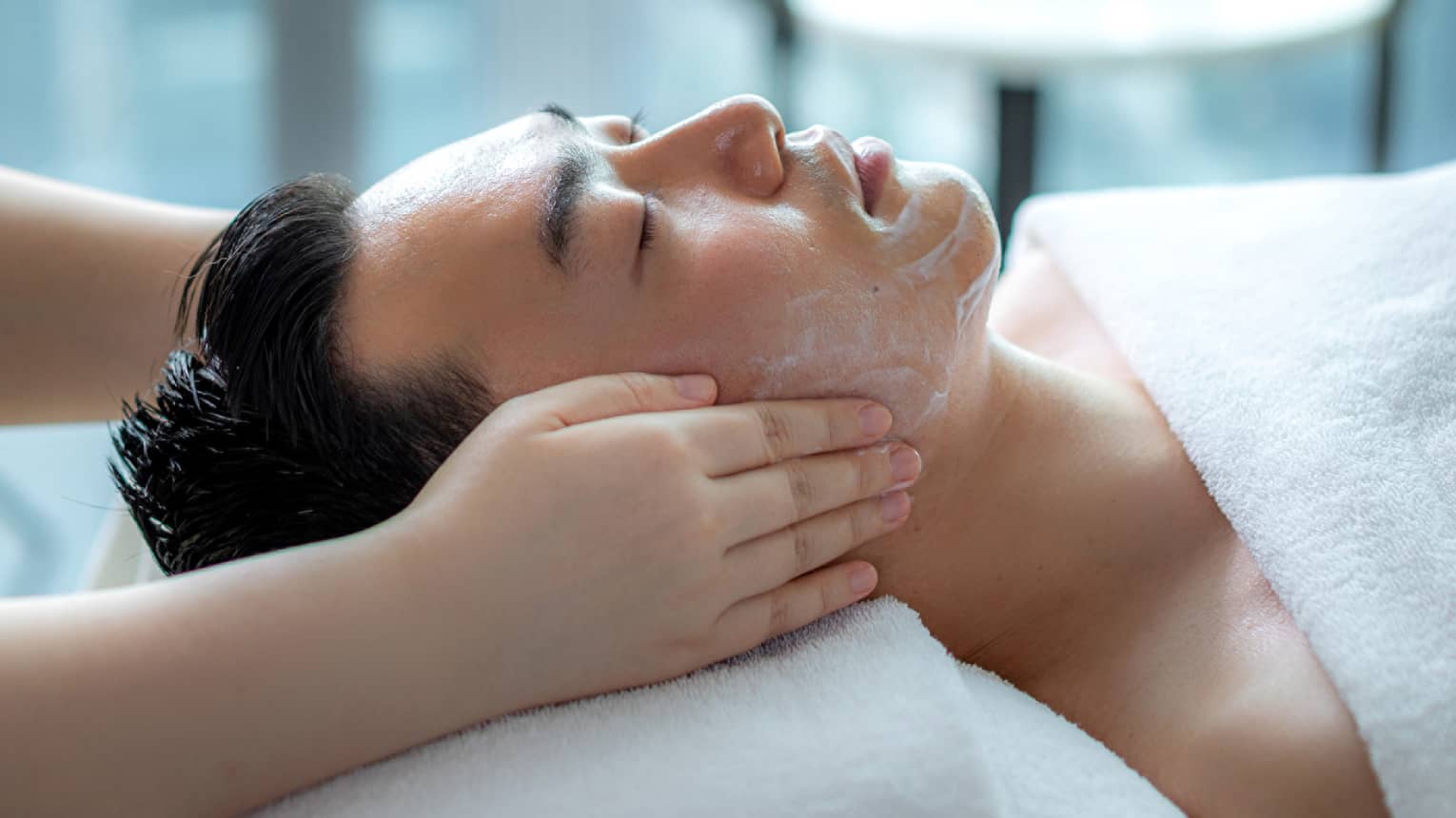 Hands rest on man's cheeks as he lays on massage table by glass window with daytime city view 