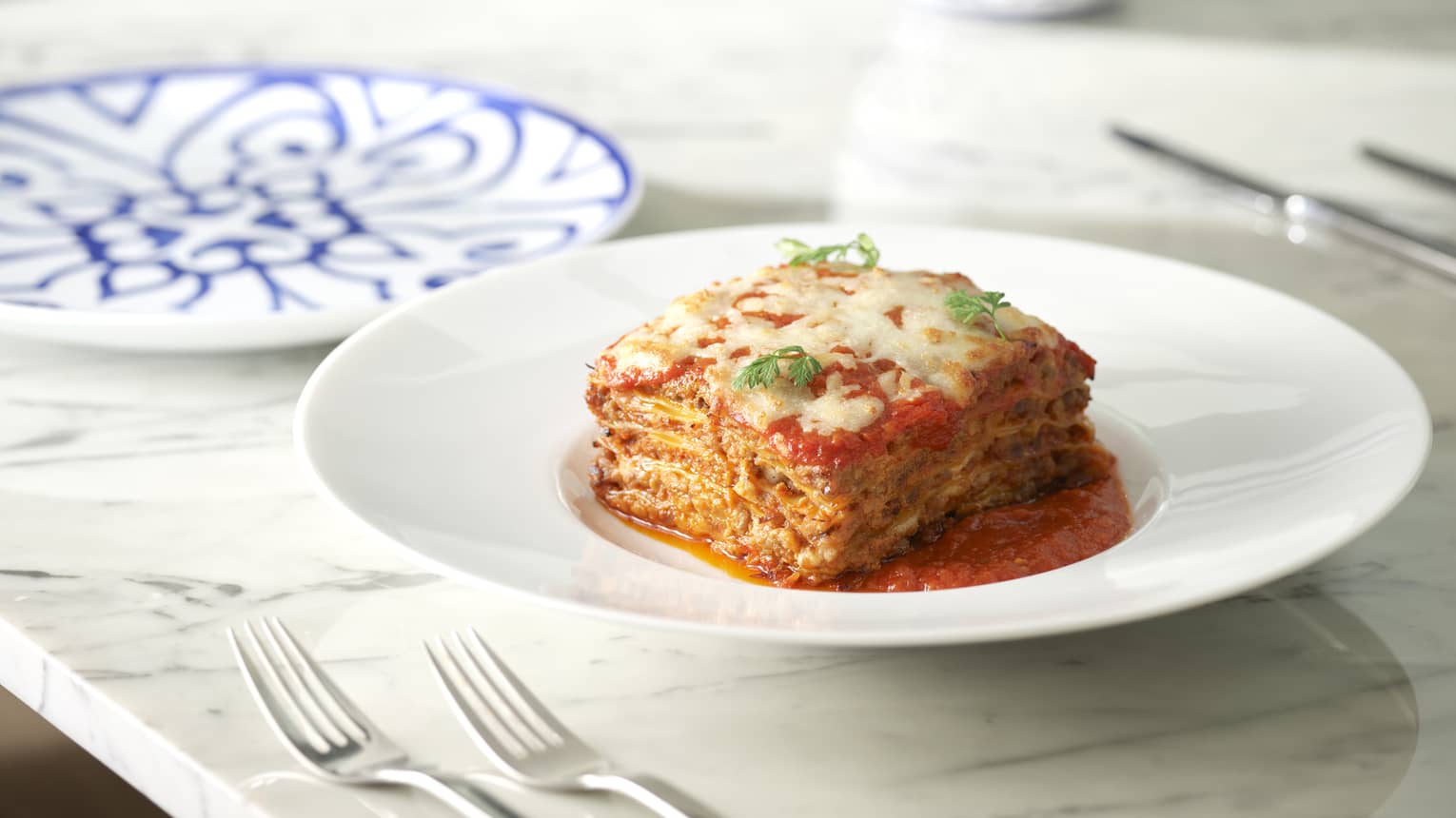 Square of lasagna in shallow white bowl on marbled table, blue-patterned china in background