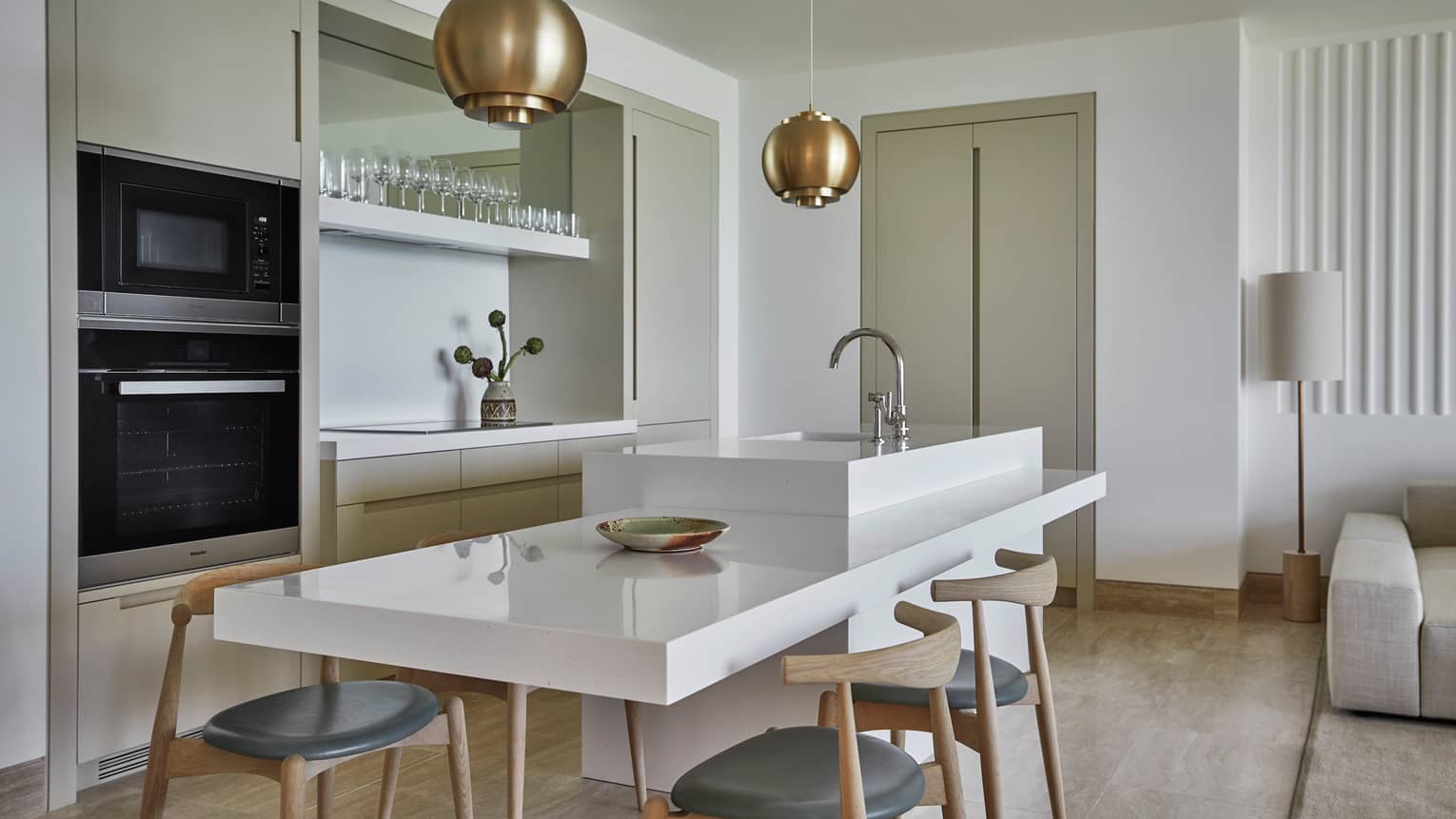 A sleek, modern in-suite kitchen in whites and beiges, featuring a large white serving island with wooden chairs and a sink.