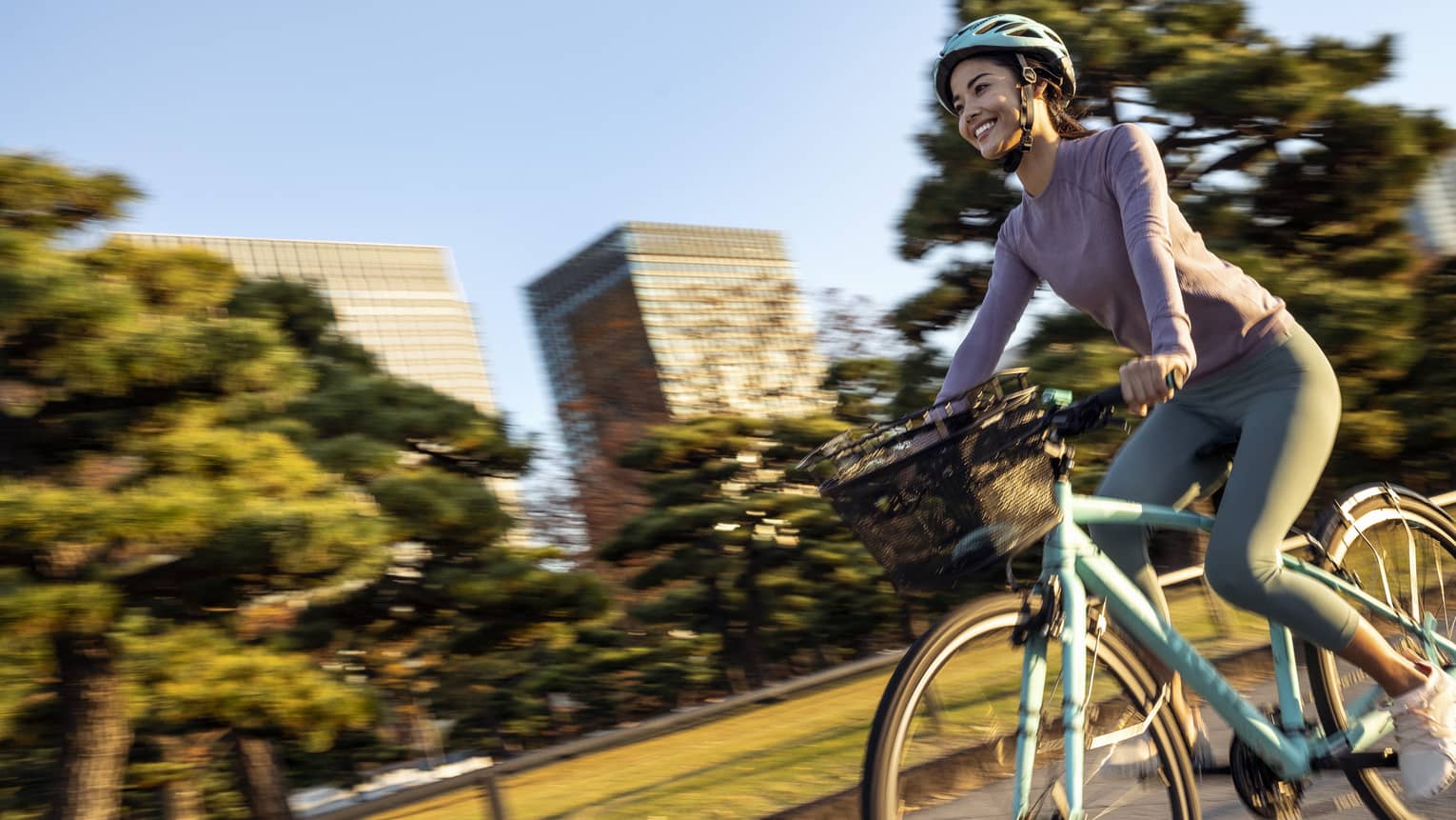 Smiling woman on teal cruiser bike with matching helmet rides by mature trees and skyscrapers of downtown Tokyo