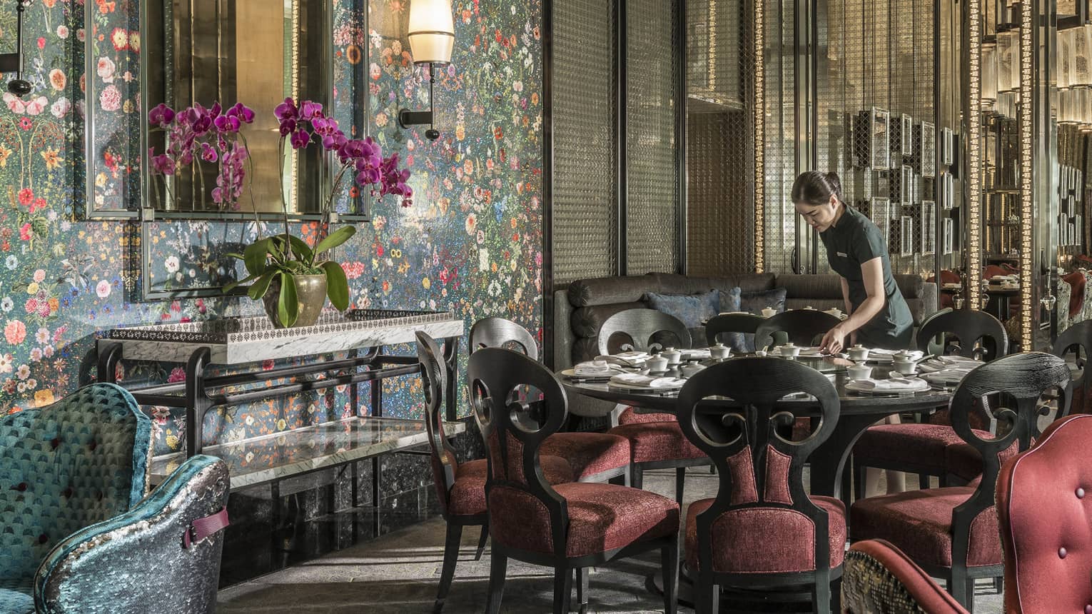 Hotel staff sets table with elegant red velvet chairs under tall mirror, floral wallpaper 