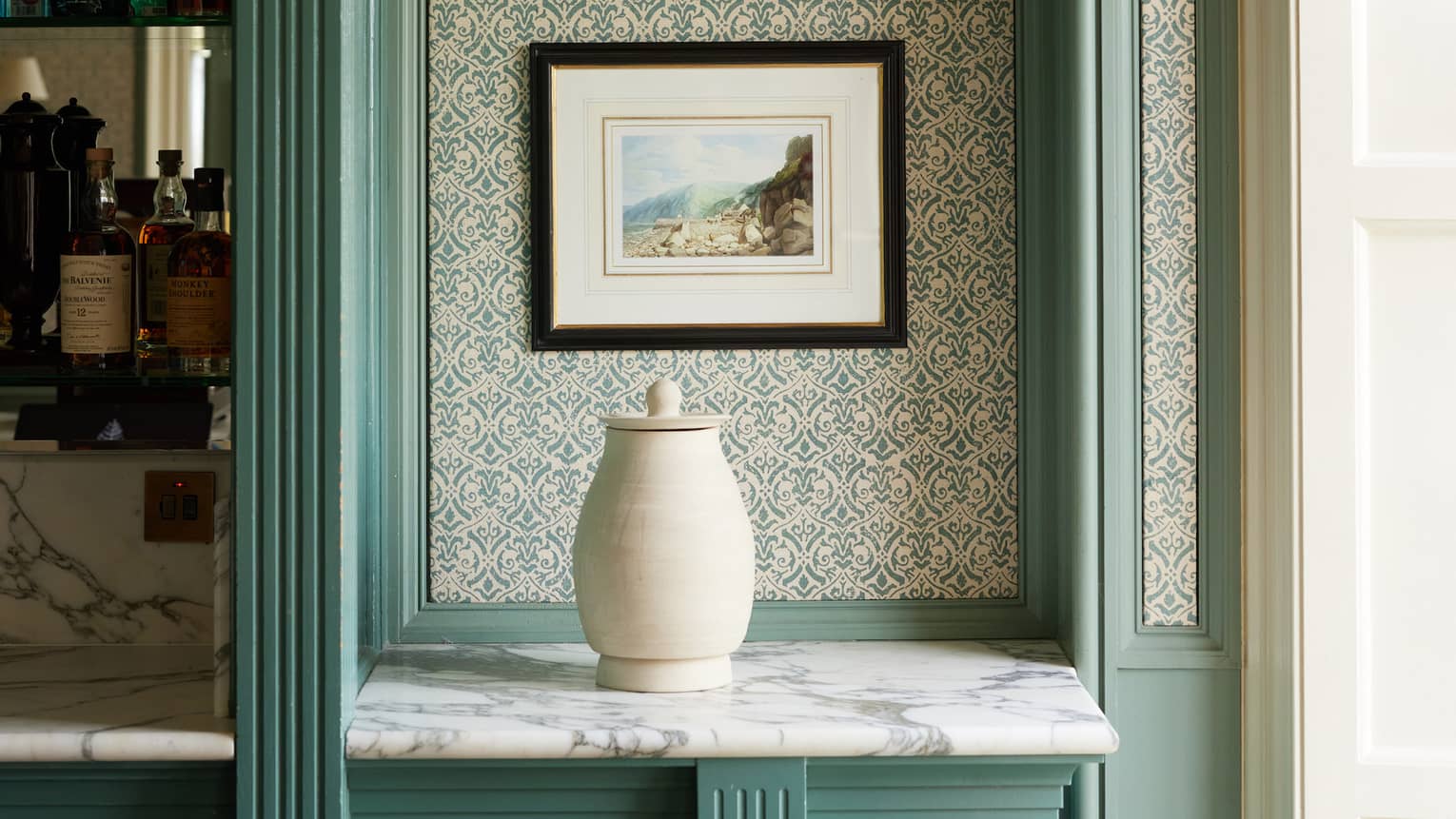Wallpapered nook with white ceramic jar on marble shelf, two stacked prints on wall and blue moulding surround