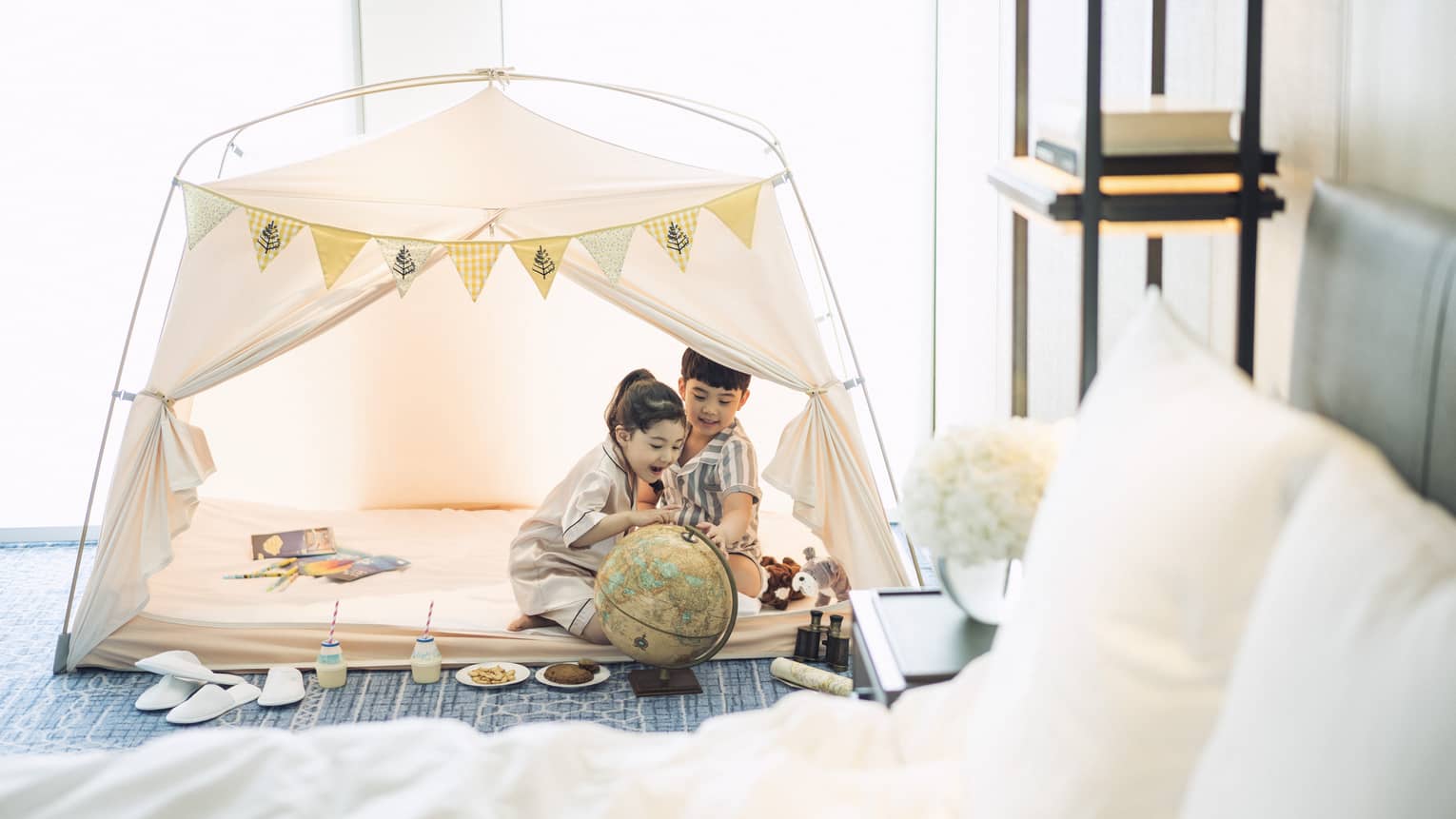 Two smiling children sit in a white play tent in a guest room at Four Seasons Hotel Seoul, with toys and snacks around them
