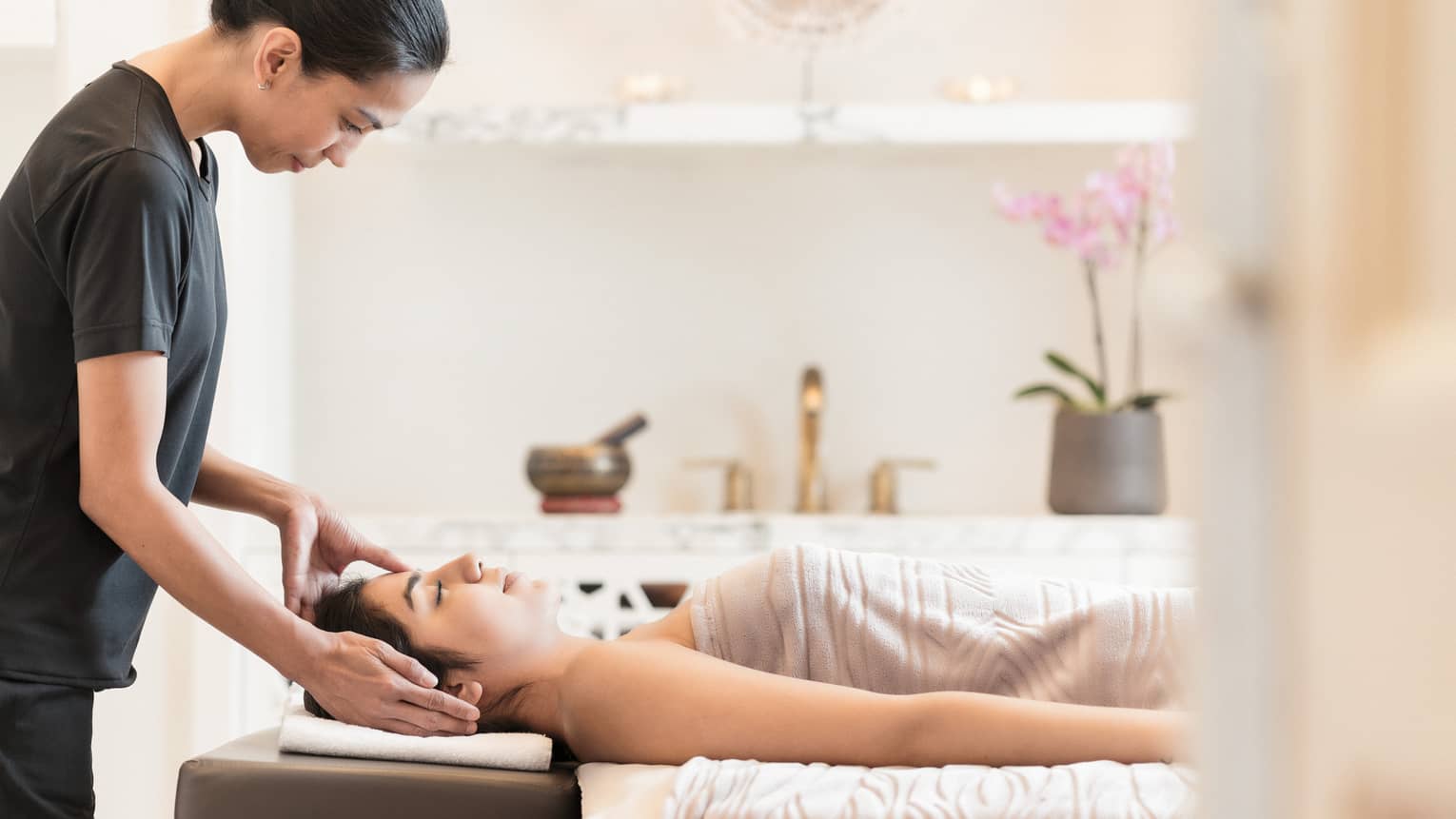 Massage therapist places hands on side of woman's face as she lays under a sheet on spa table