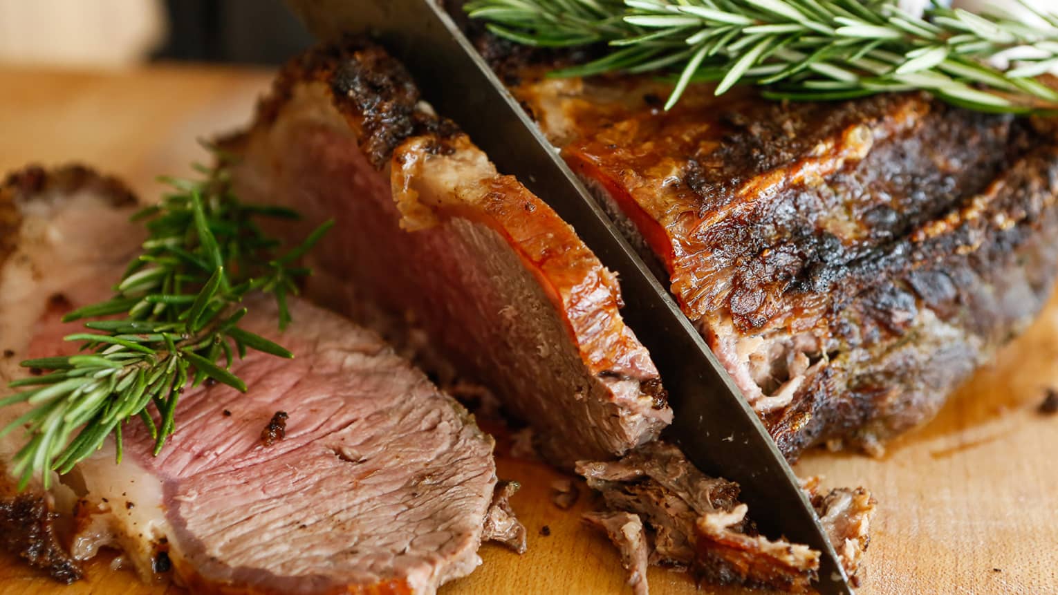 Close up of knife carving slice of pork roast topped with fresh rosemary sprigs