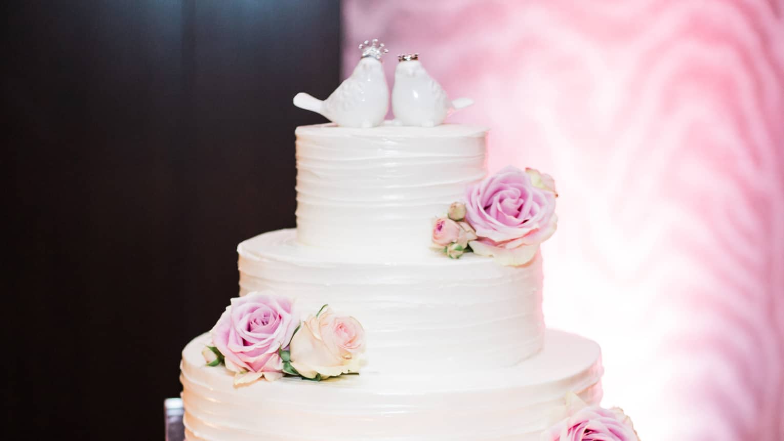 Three tiered white wedding cake decorated with fresh pink roses, two ceramic birds 