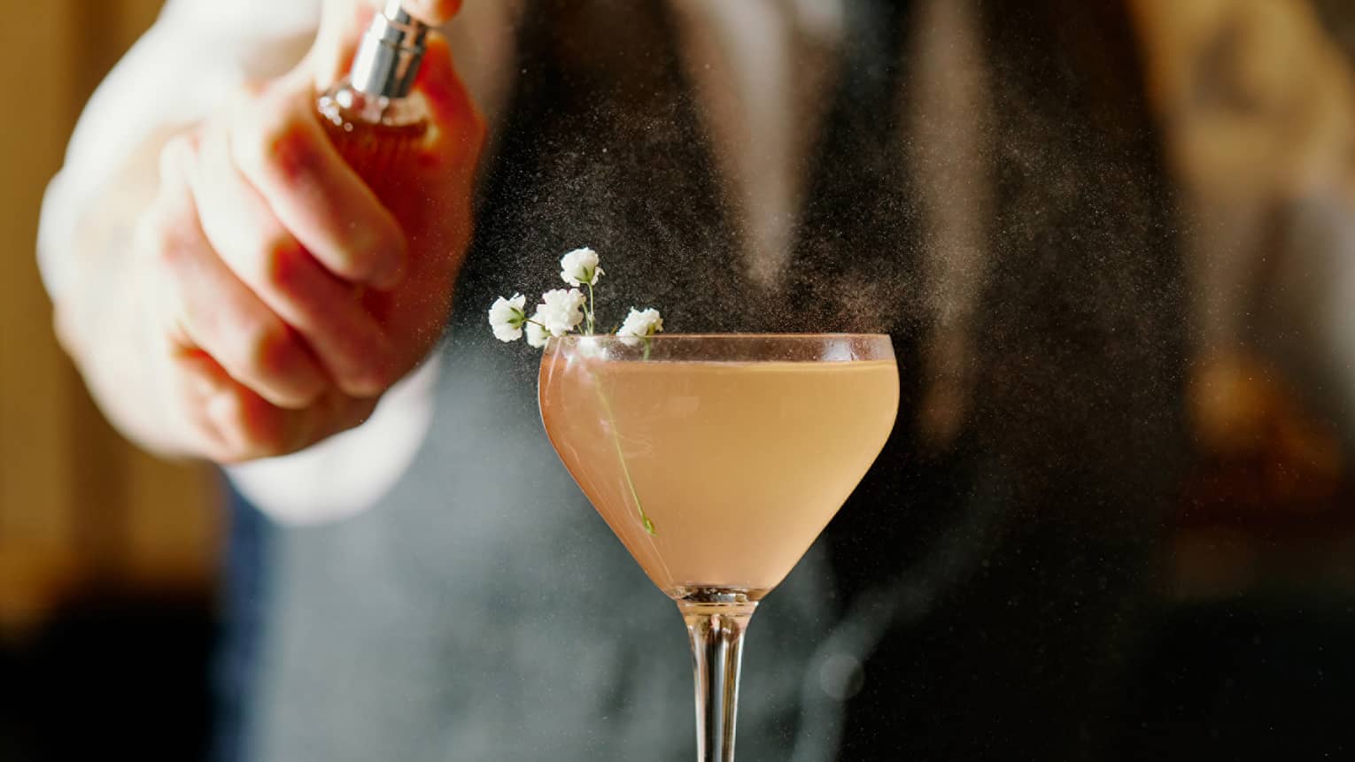 A light yellow cocktail with small white flowers on being sprayed by a small bottle by a bartender.
