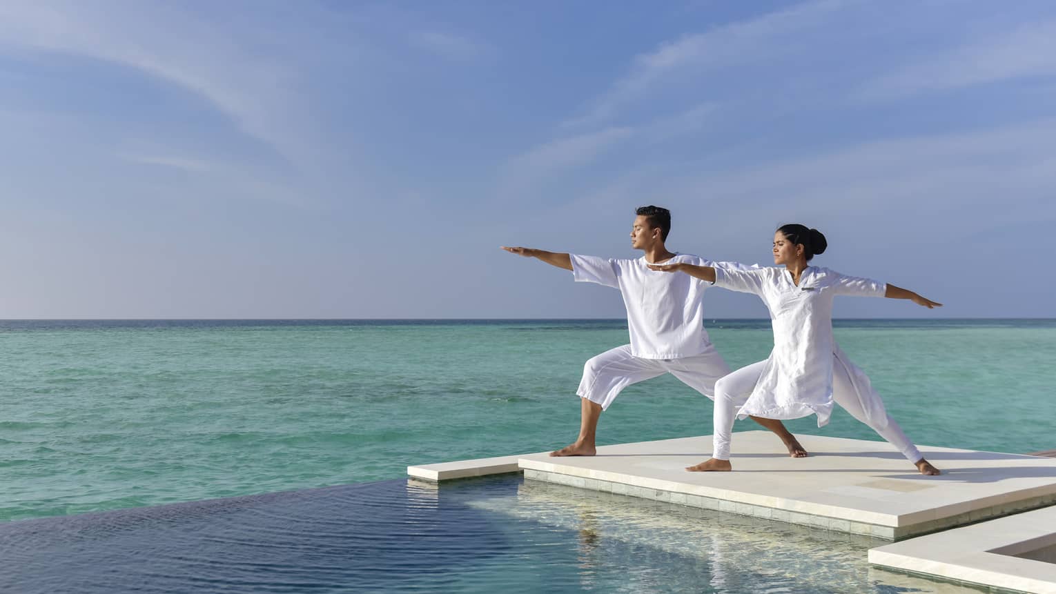 A couple dressed in all white does yoga on a white dock over the sea
