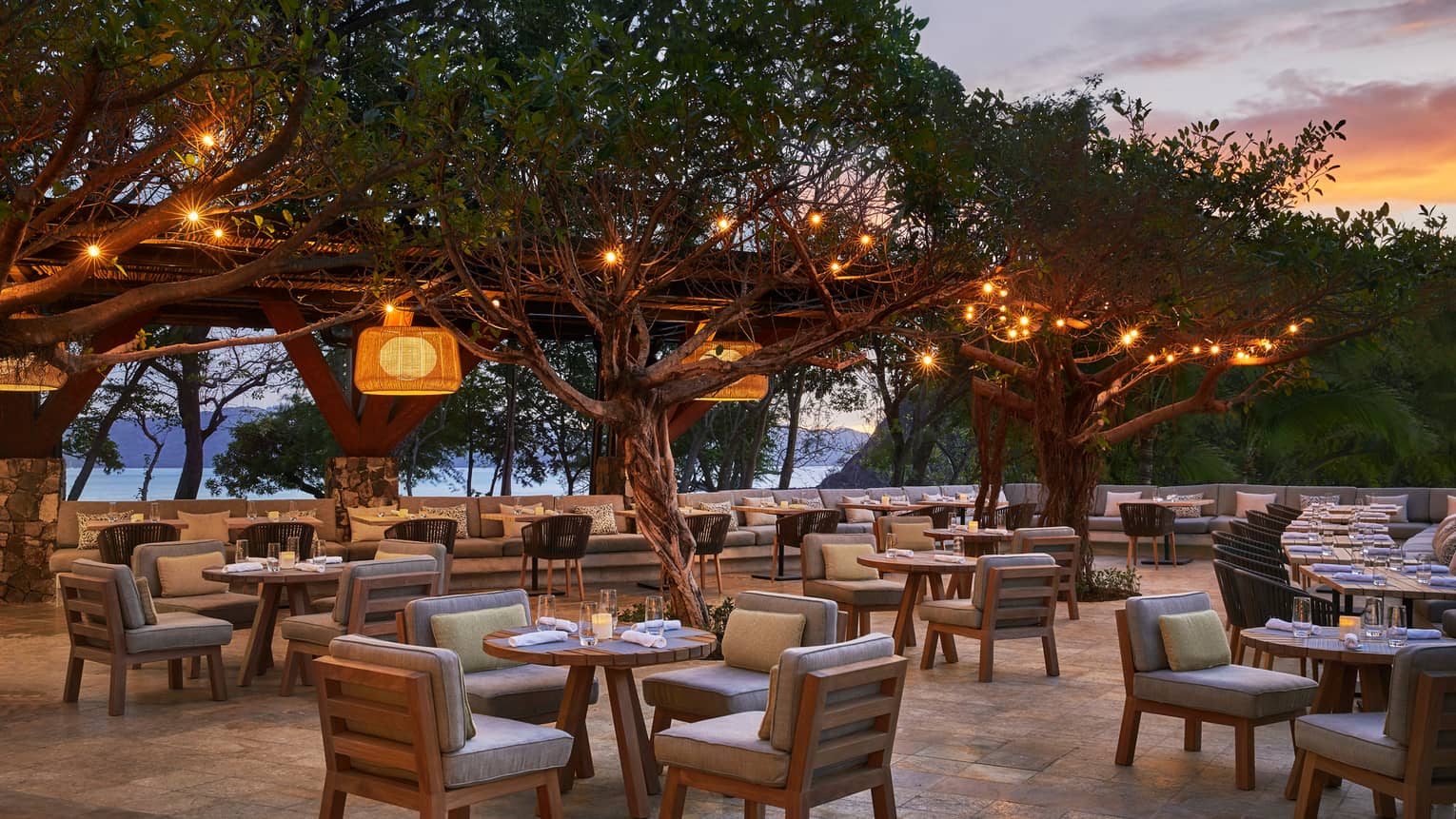 Patio tables, chairs with plush cushions on patio under trees with hanging lights, at sunset