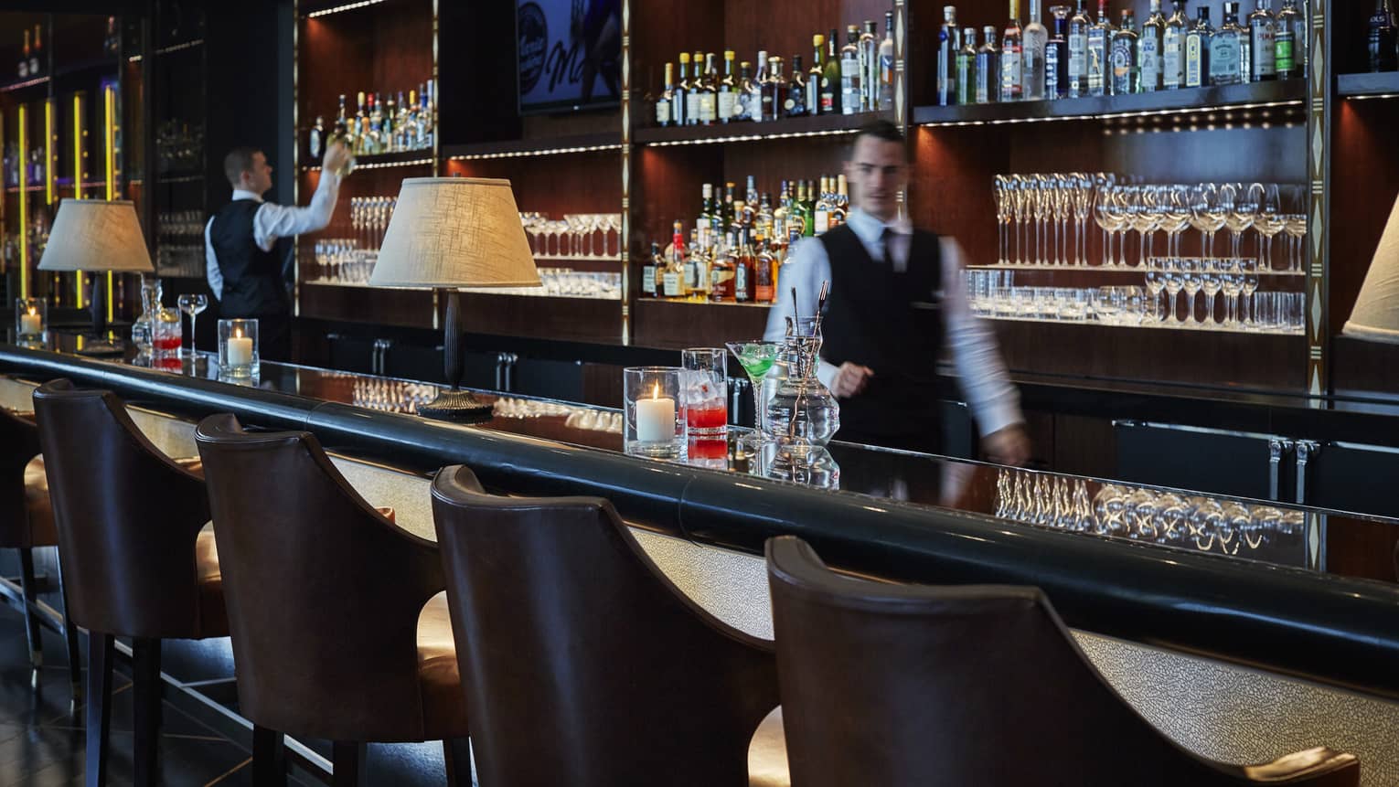 Bartender in black vest, white shirt, walks behind dimly-lit bar lined with leather bucket stools