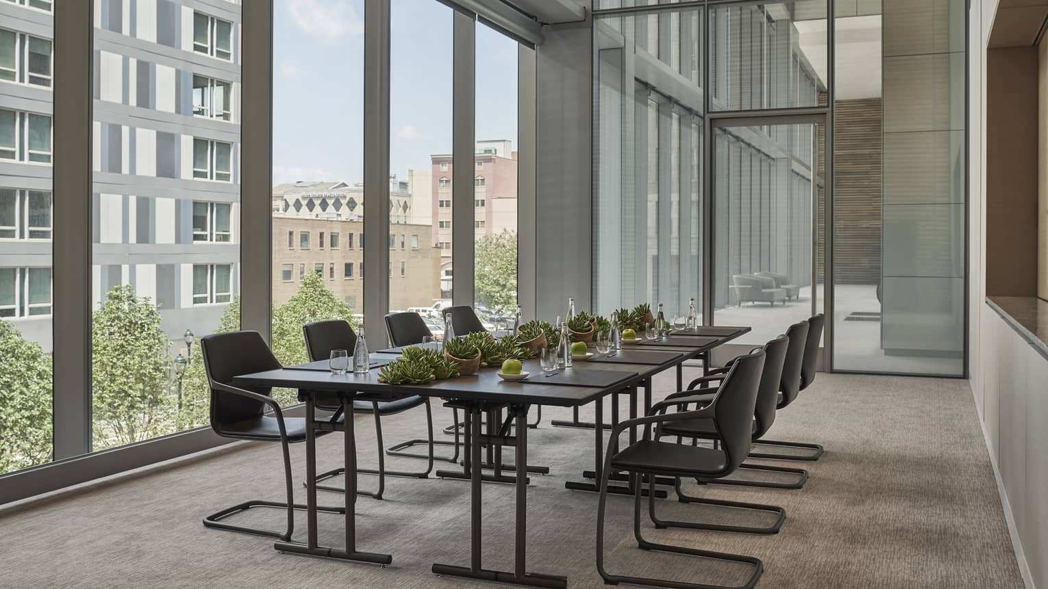 A boardroom table with many chairs next to a wall covered in large windows looking out at a city.