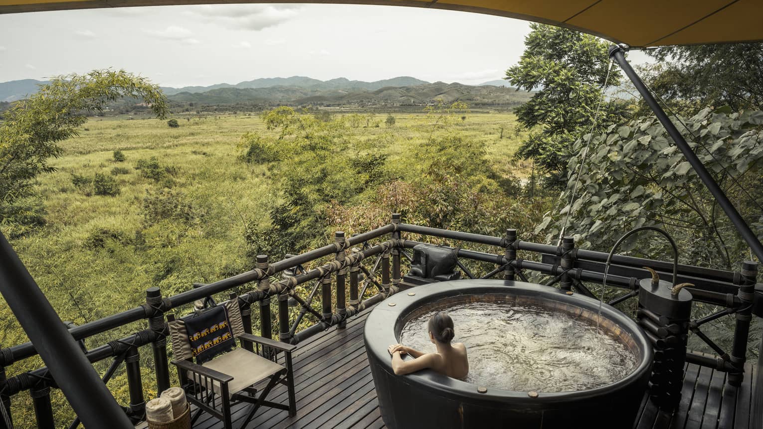 Woman wades in large round hot tub on wood deck overlooking green field, mountains