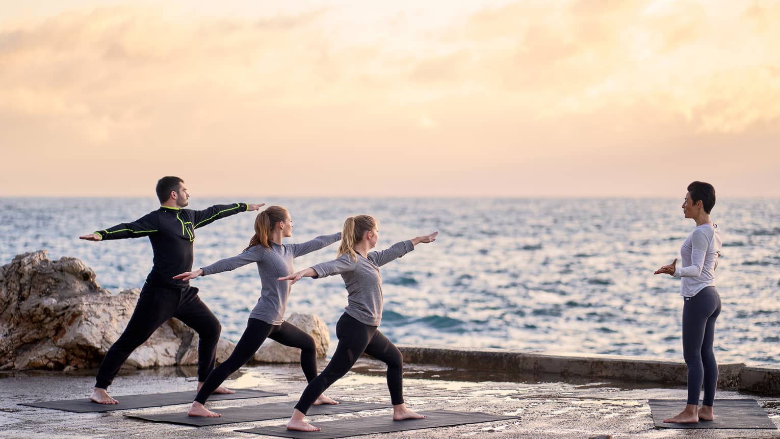 Three people in warrior yoga pose and coach standing at sea's edge at sunrise