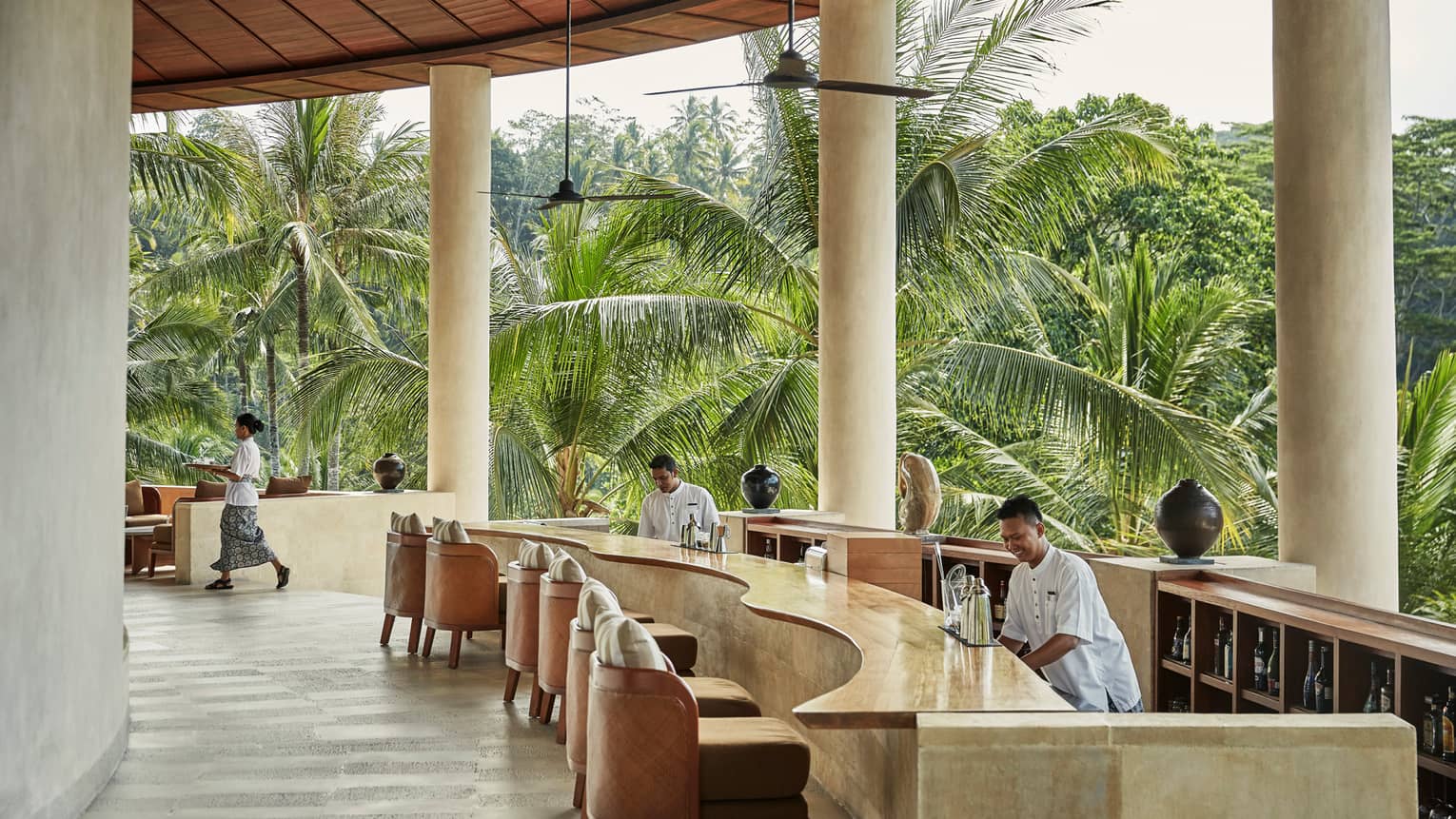 Smiling bartenders behind long wood-and-stone bar under white pillars of resort balcony, palm trees in background