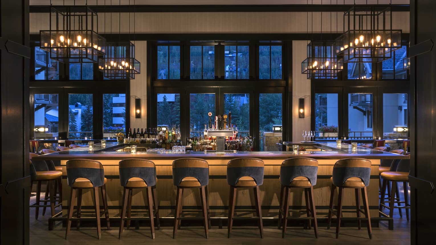 Dimly-lit bar at dusk lined with retro-style wood stools, lantern chandeliers above, large windows