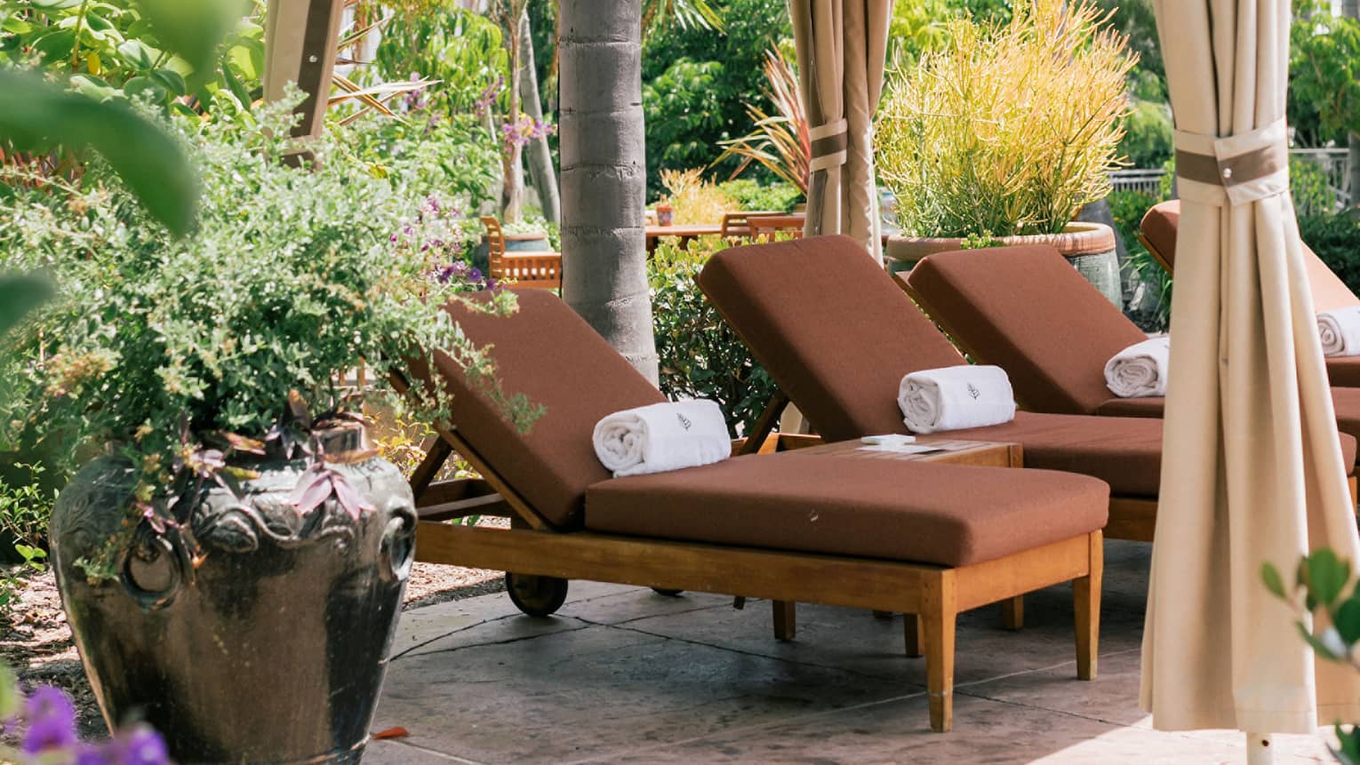 A poolside cabana with brown lounge chairs and plants and flowers surrounding it.