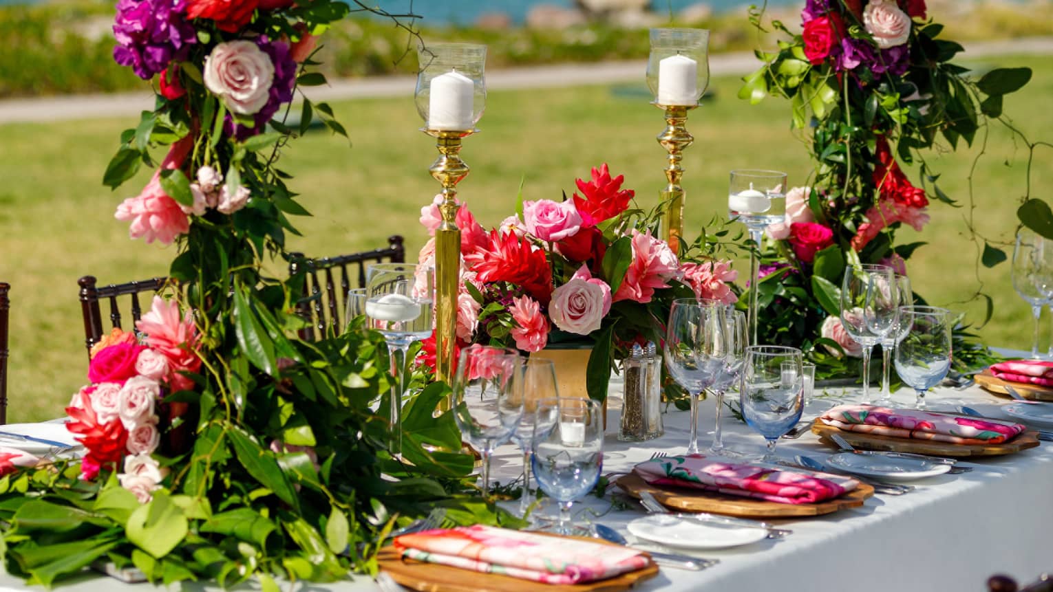 Angled colourfully set dining table, tall pillar candles, large circular floral centrepiece, water views