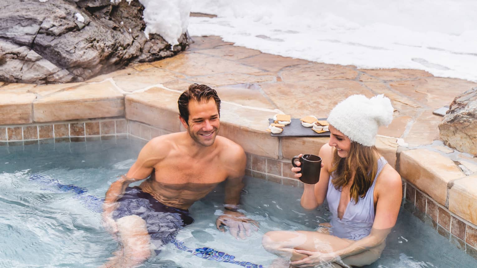 A man and woman in lounging in a heated pool surrounded by snow.