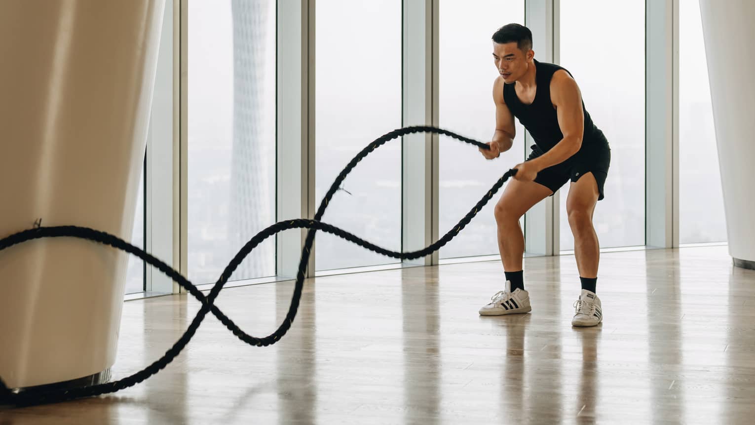 Front view of a well-toned guest moving battle ropes in a wave pattern on a pale wood floor next to floor-to-ceiling windows.