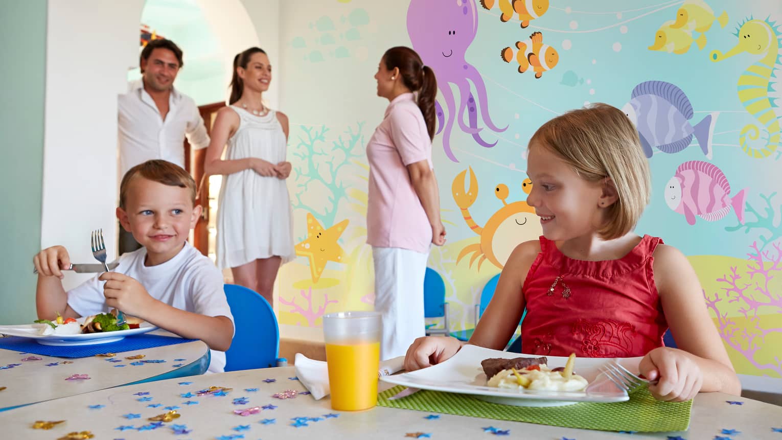 Two smiling young children at table with plates of lunch, glass of orange juice, parents and staff in front of colourful sea mural
