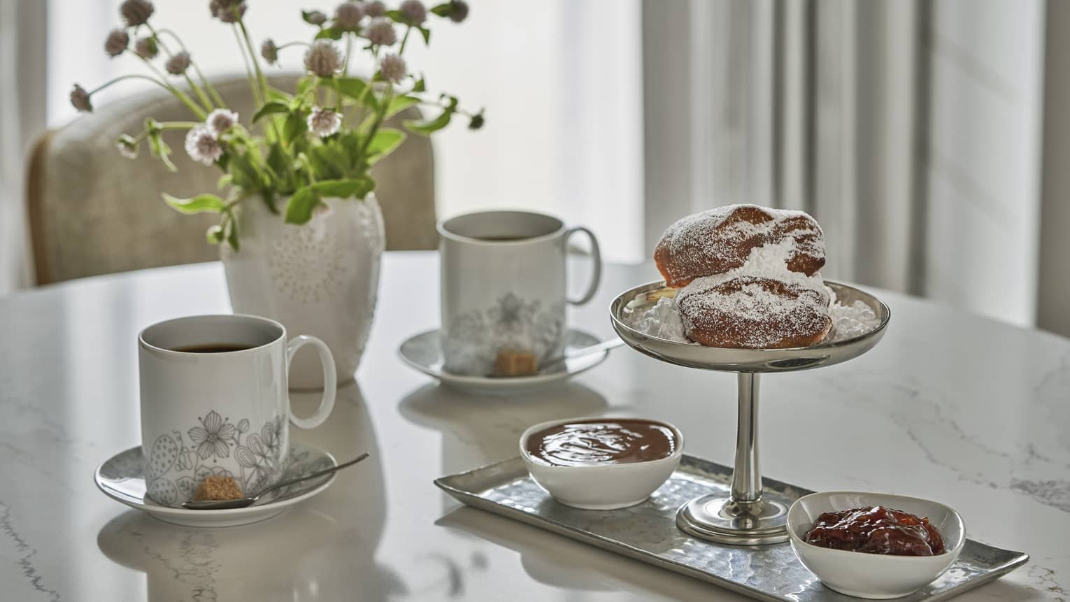 Marble-topped table with a silver tray of beignets and condiments, two mugs of coffee and a vase of fresh wildflowers