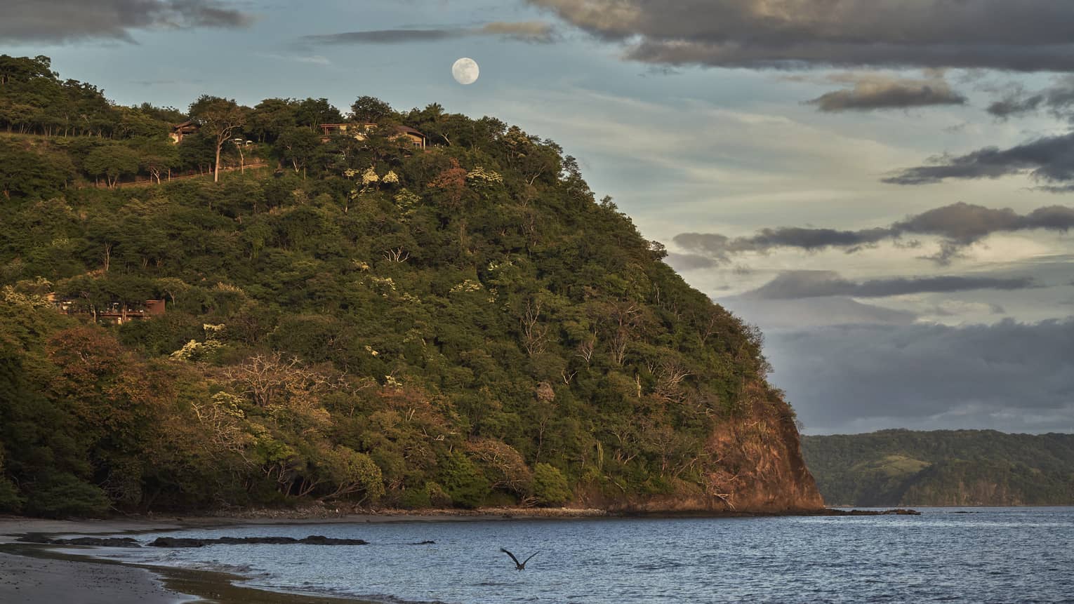 A full moon rises above a seaside cliff in a blue, daytime sky