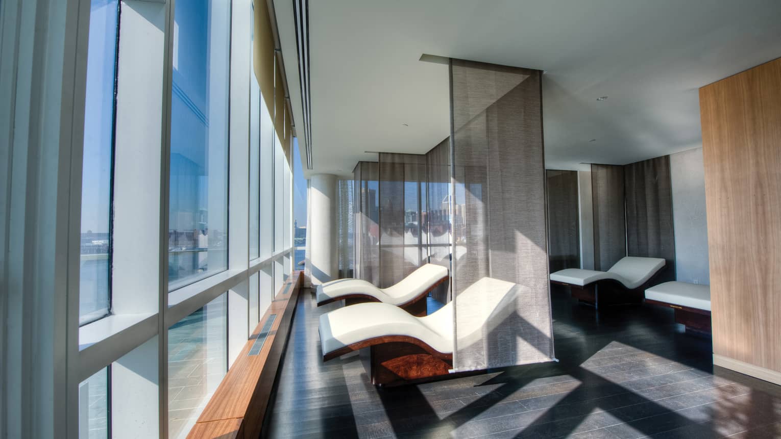 Two curved white spa chairs divided by sheer panel curtains in front of floor-to-ceiling windows