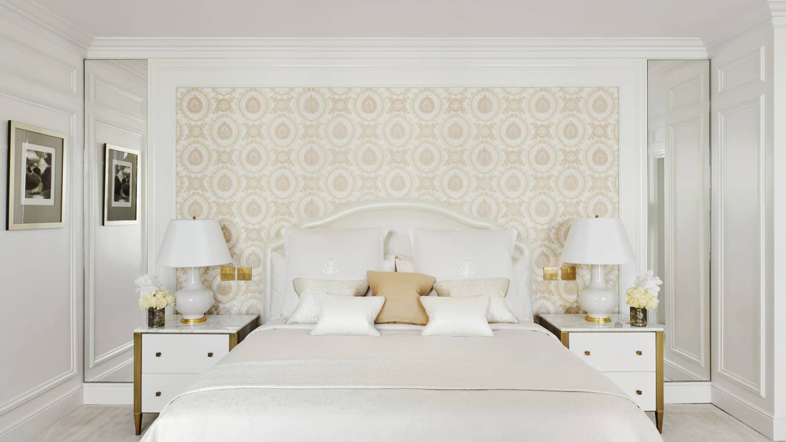 All-white bed flanked by white nightstands and lamps against beige wallpapered wall 