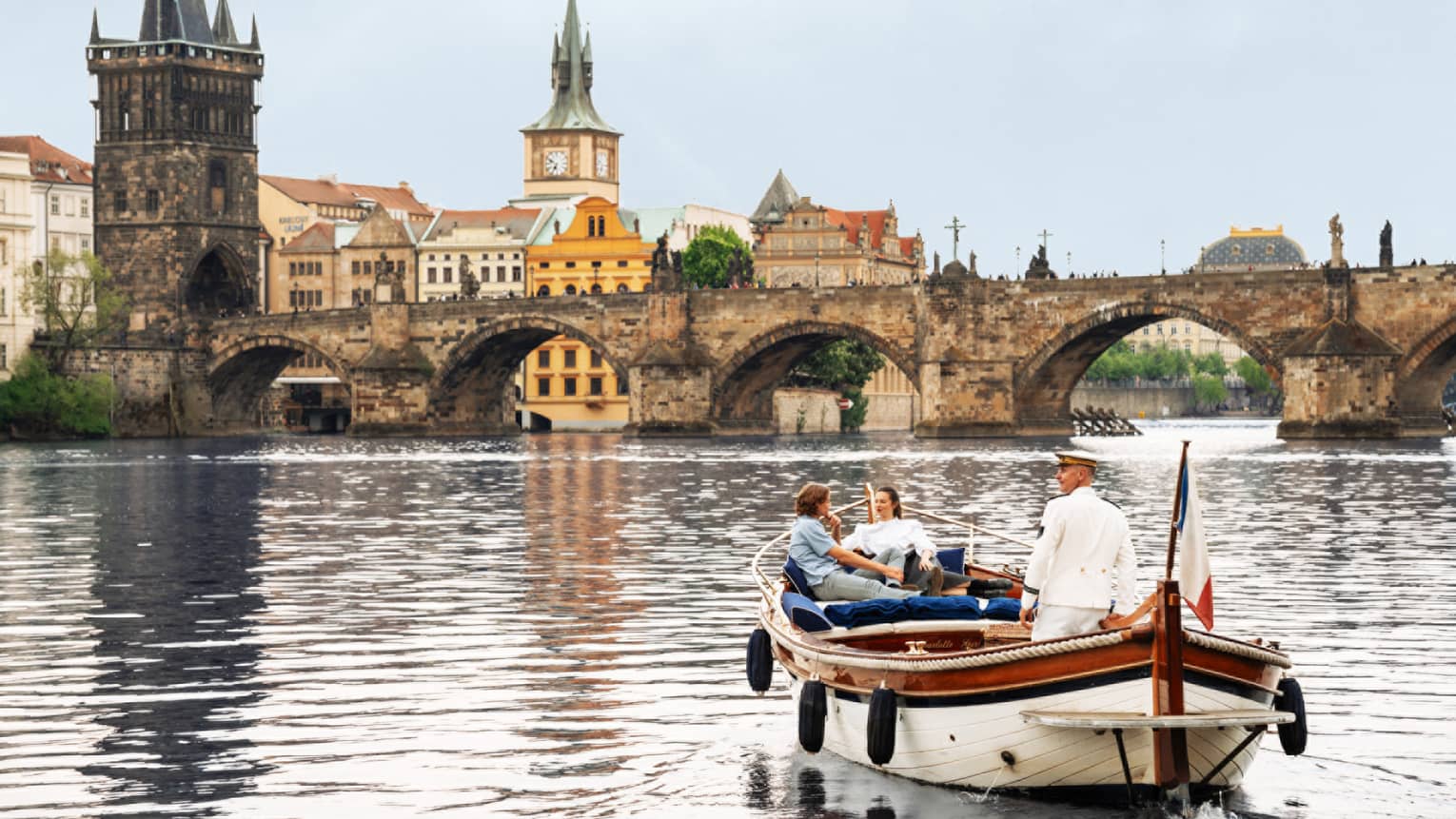 Couple with captain in wooden boat on Vltava River with bridge and buildings in backdrop