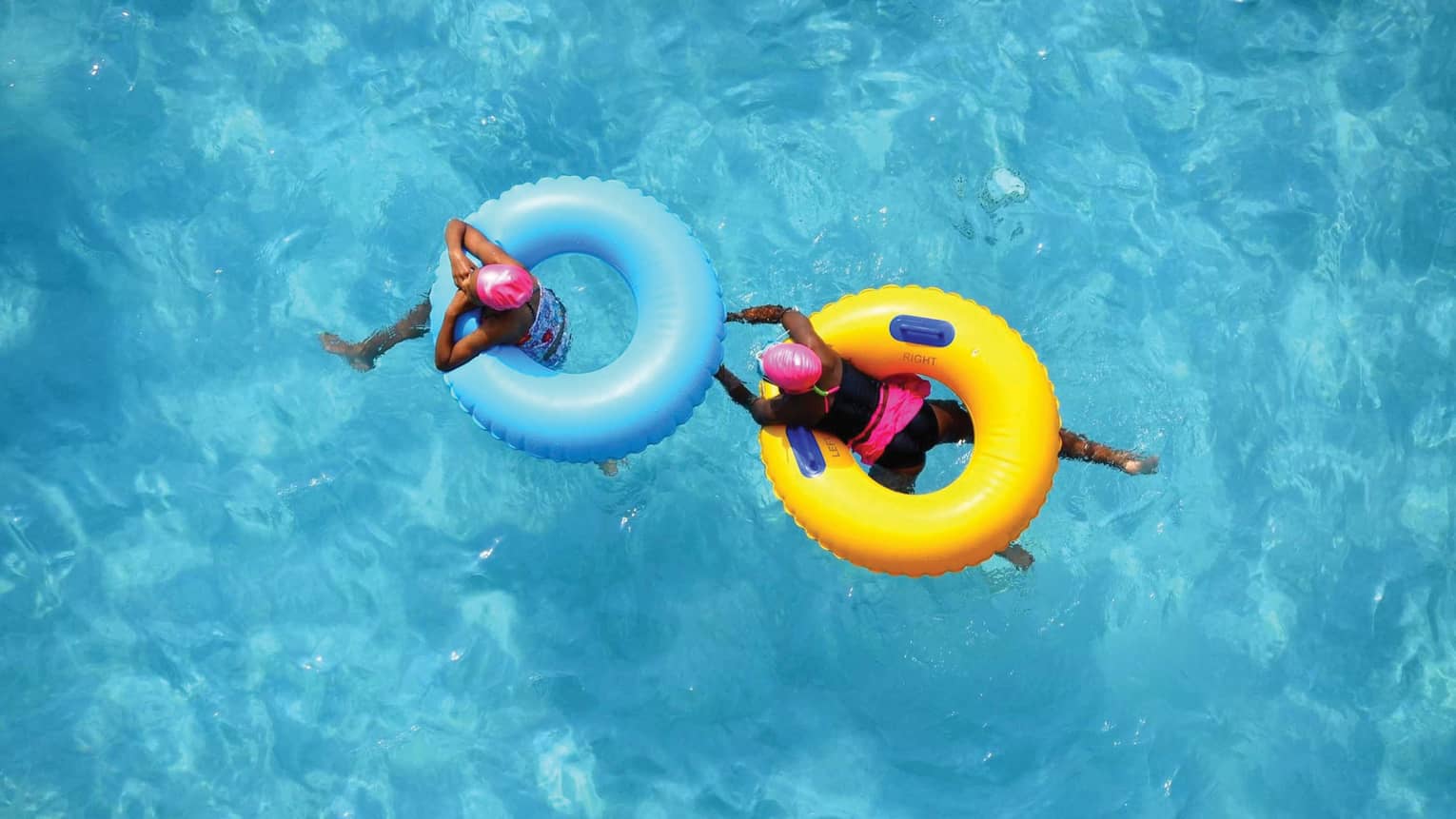 Aerial view of two swimmers in pink caps floating in blue and yellow inner tubes 