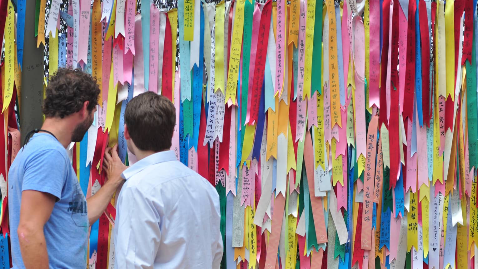 Two men looking at barbwire fence entirely covered with colourful ribbons with Korean text expressing wishes for world peace.