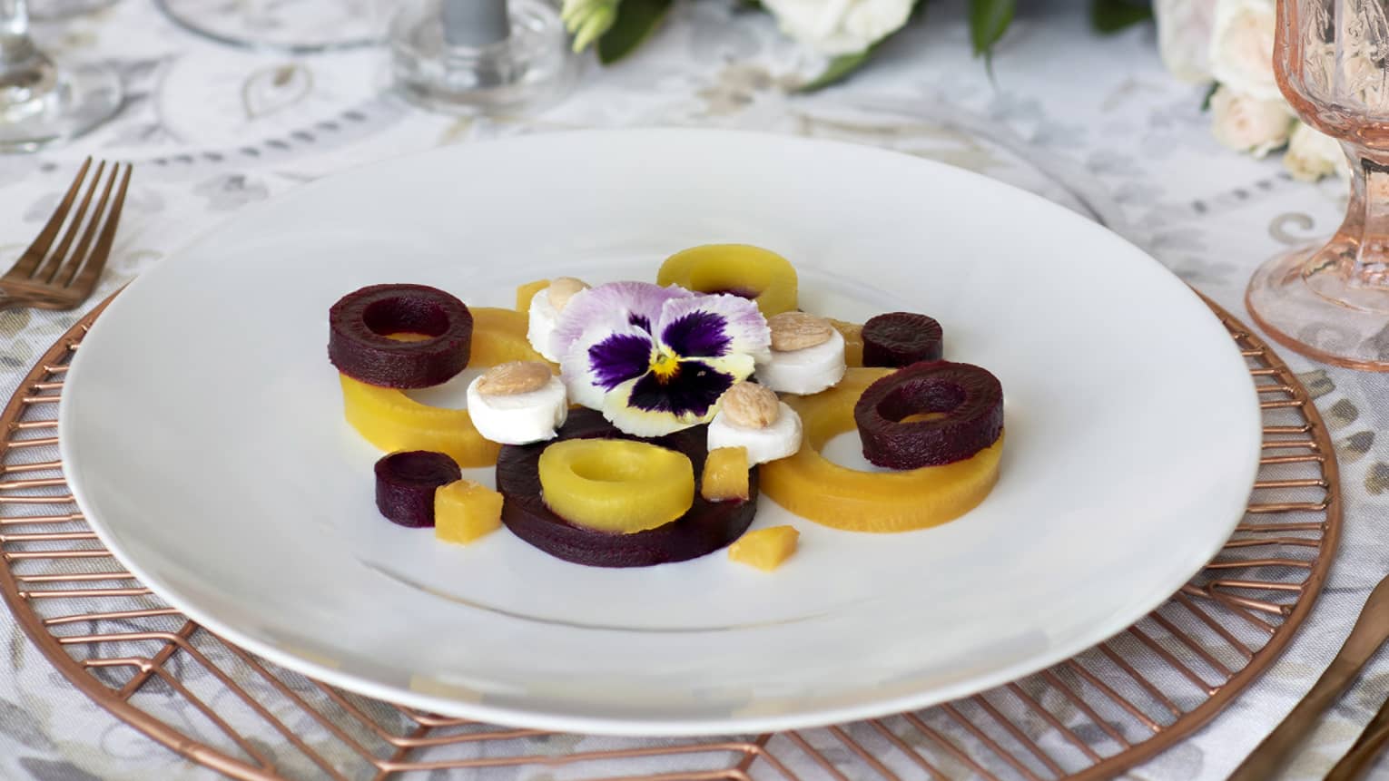 White roses behind plate with round beets, goat cheese, nuts and flower