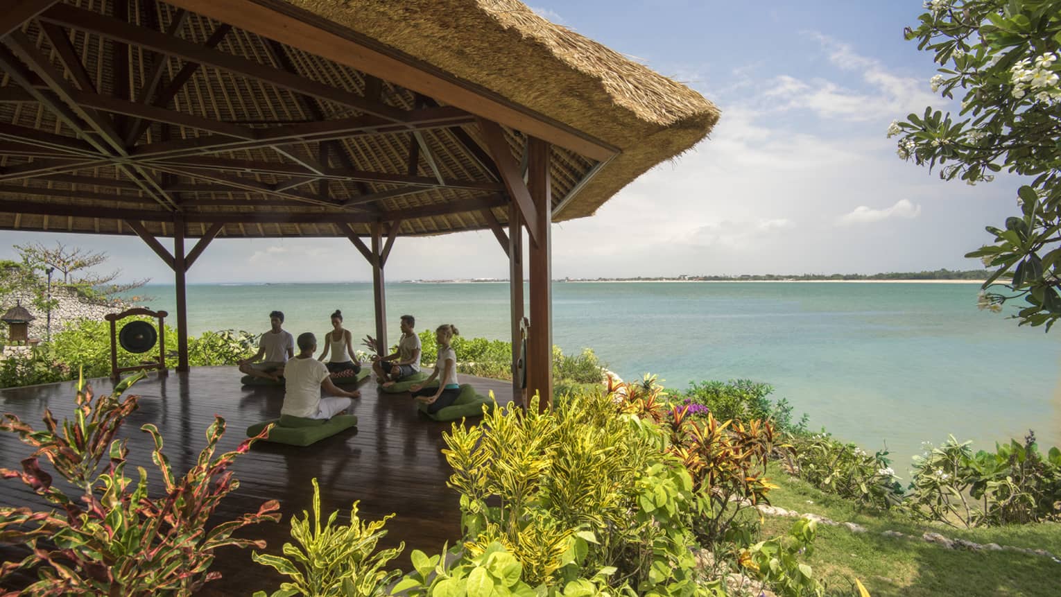 Five people sit cross-legged meditating on green cushions under thatched-roof gazebo by ocean