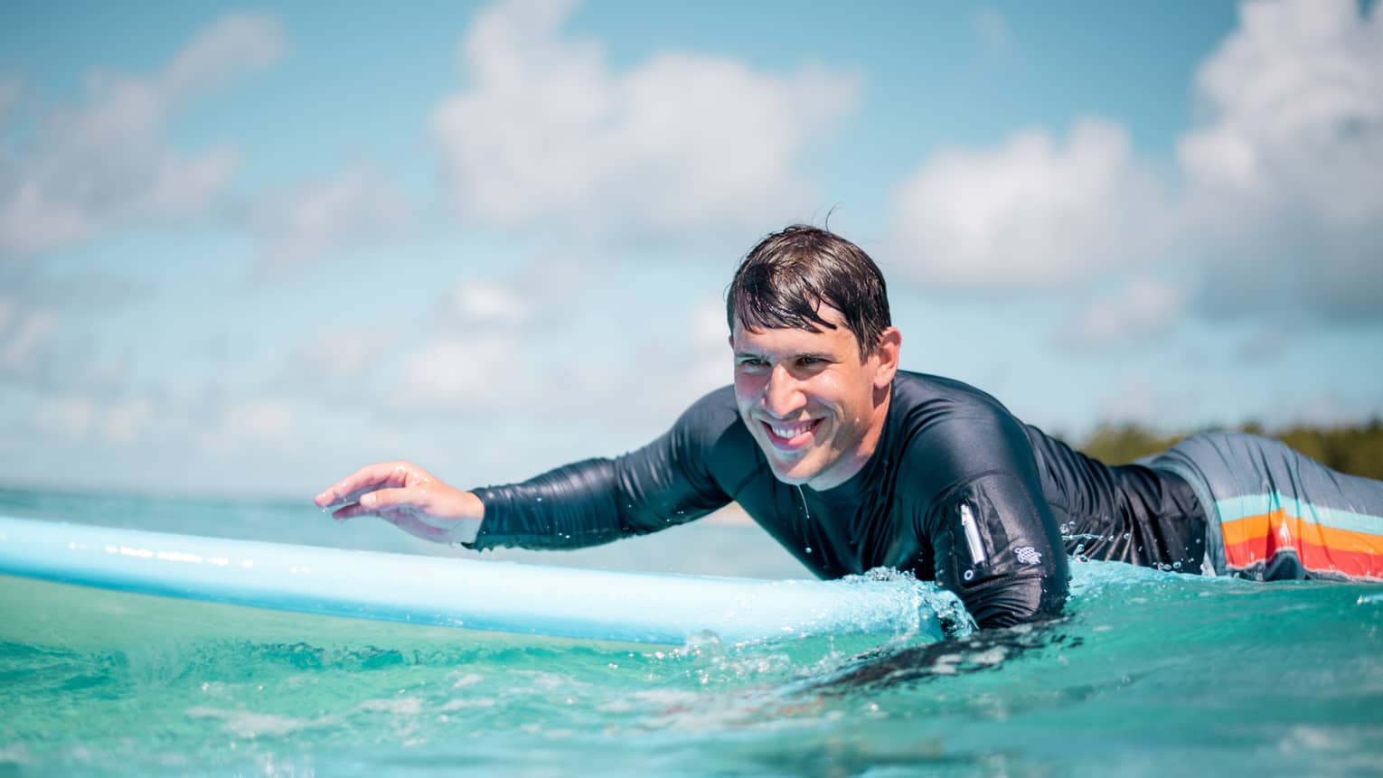 Man in rash guard smiling while paddling on blue surfboard in blue-green water