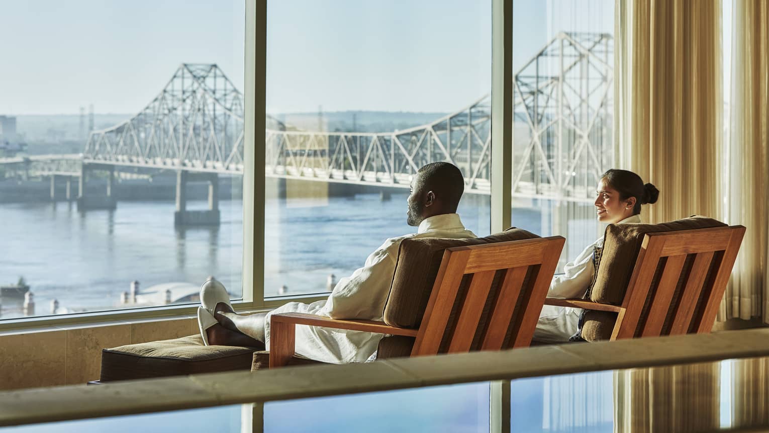 Couple lounge in chairs by large windows overlooking water, St. Louis bridge