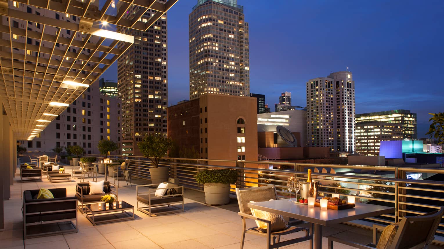 Large rooftop patio at night with candle-lit tables, champagne on ice, black cushioned chairs, city lights