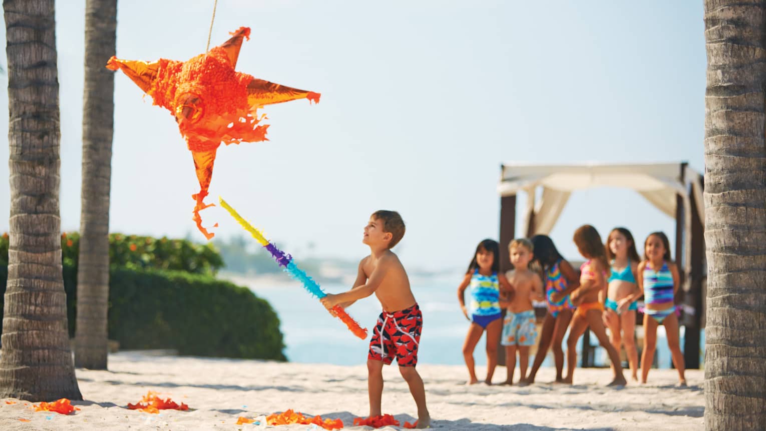 On a beach, a child swings a bat wrapped in vibrant crepe paper at a big orange star piñata; other children watch excitedly. 