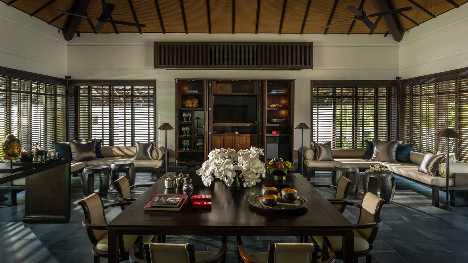 Large dining table with flowers in middle of room lined with sofas, windows, shutters